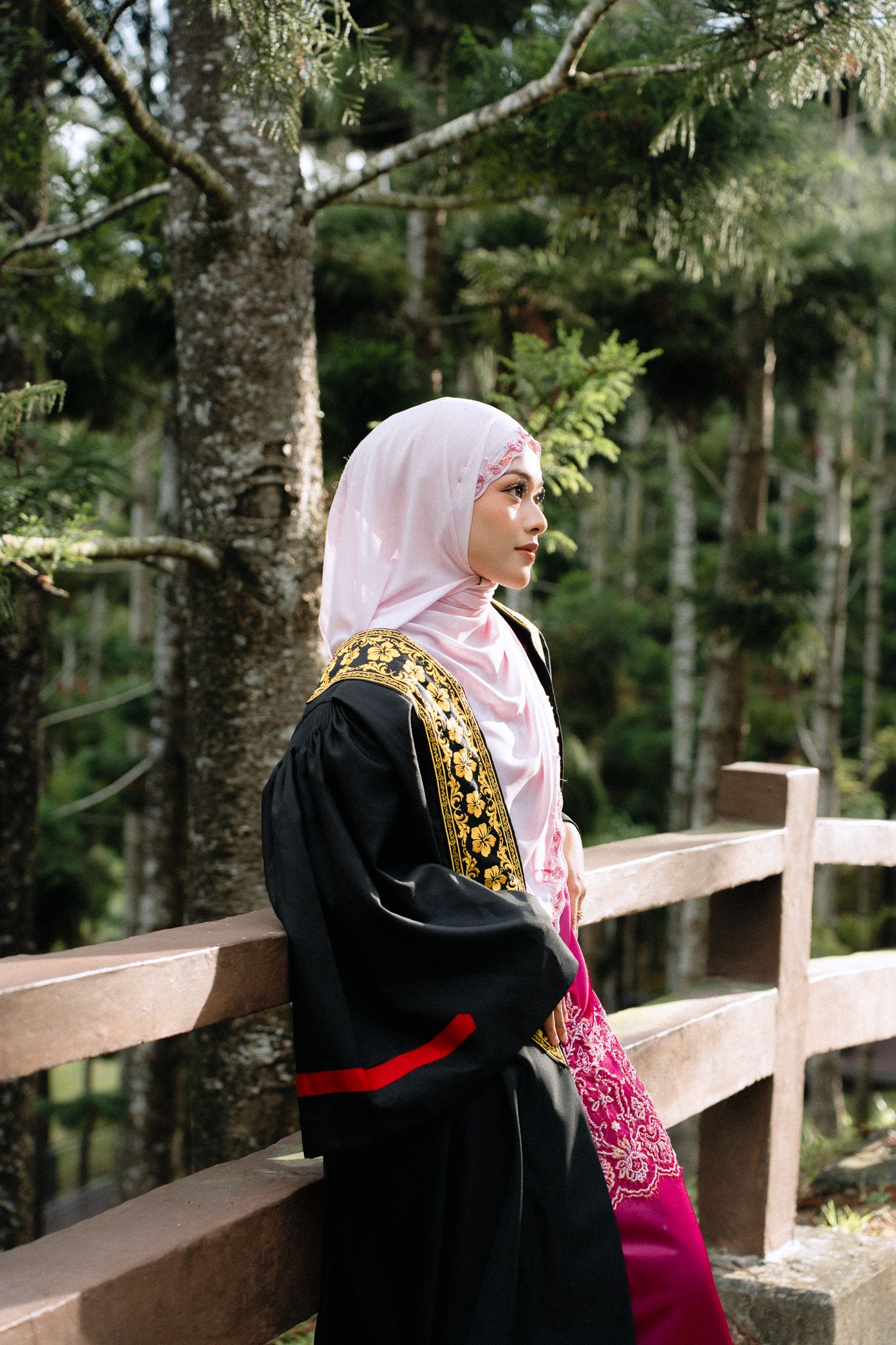A Muslim woman in a black abaya and pink hijab stands by a wooden railing surrounded by tall trees in a forest.