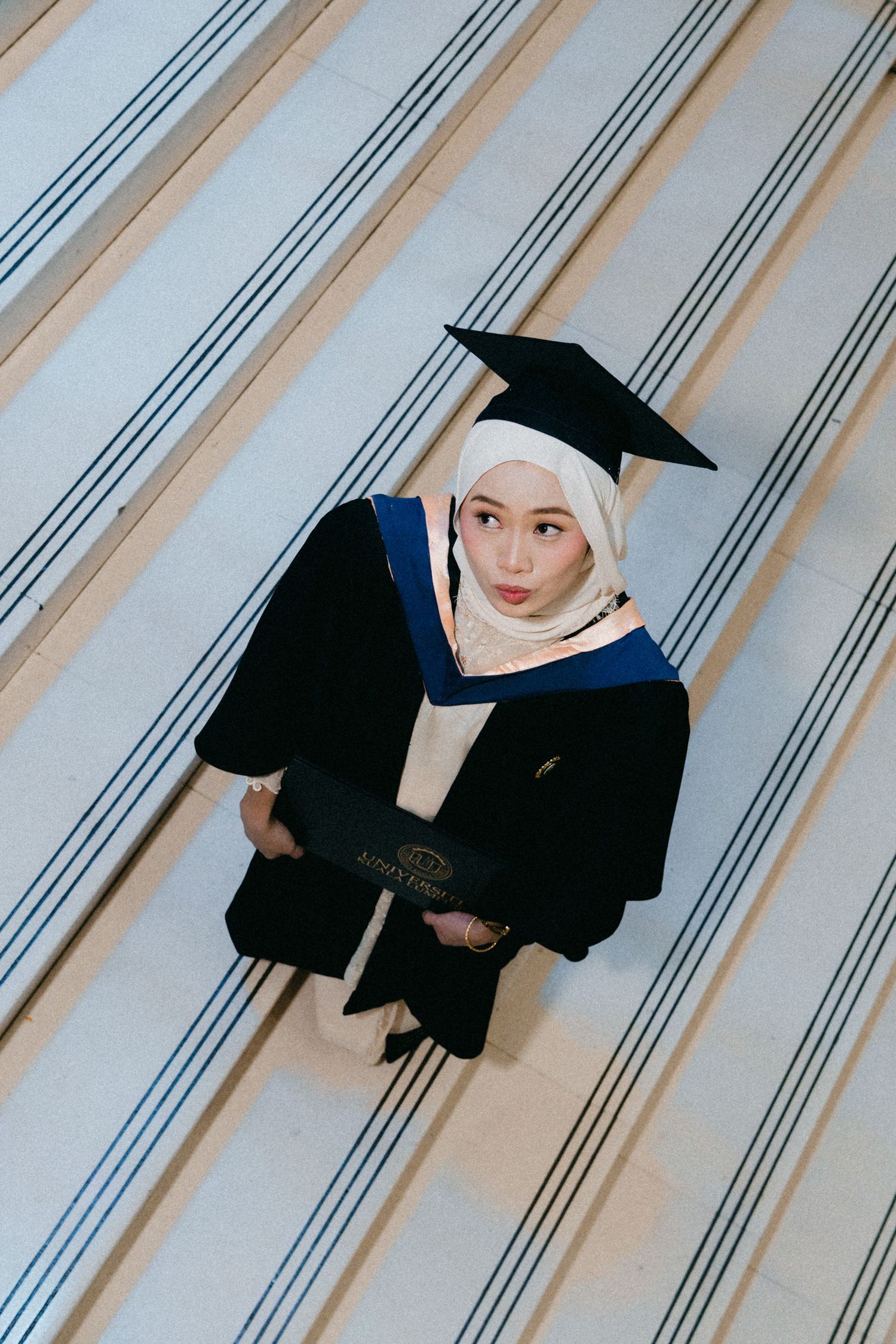 Graduate in black academic gown and cap poses on striped carpeted stairs.