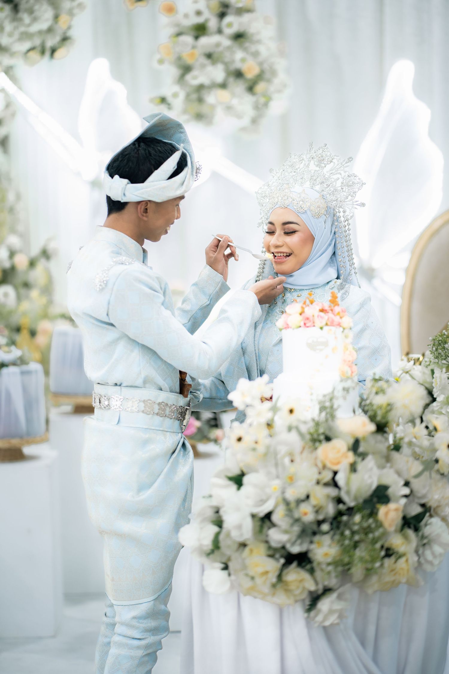 A tender moment between newlyweds in white and light blue traditional Muslim wedding attire surrounded by floral decor.