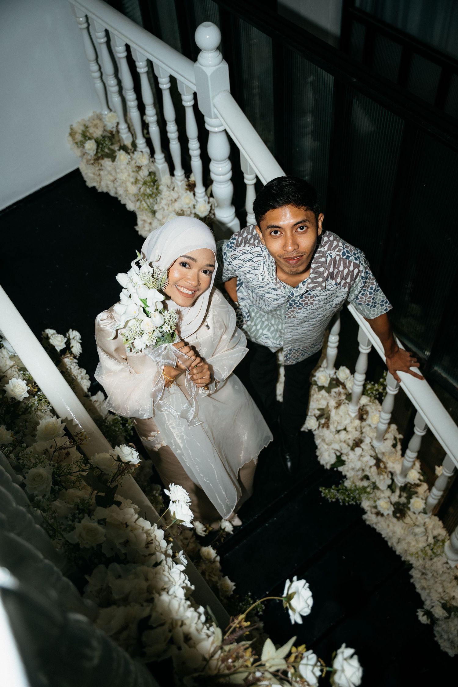 A couple stands on decorated stairs with white flowers during what appears to be their wedding celebration.