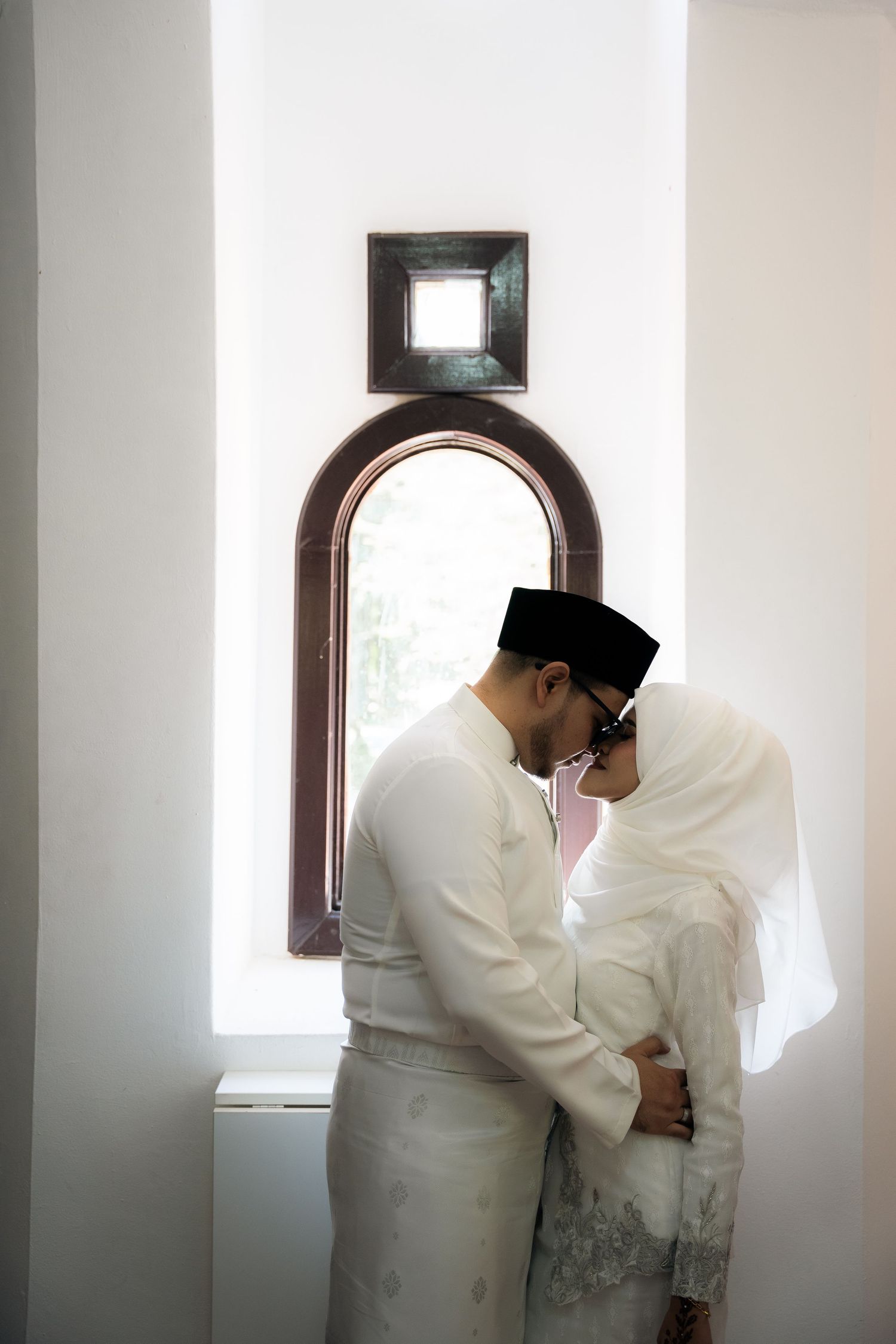 A couple in traditional white attire share an intimate moment by a window with arched wooden frame.