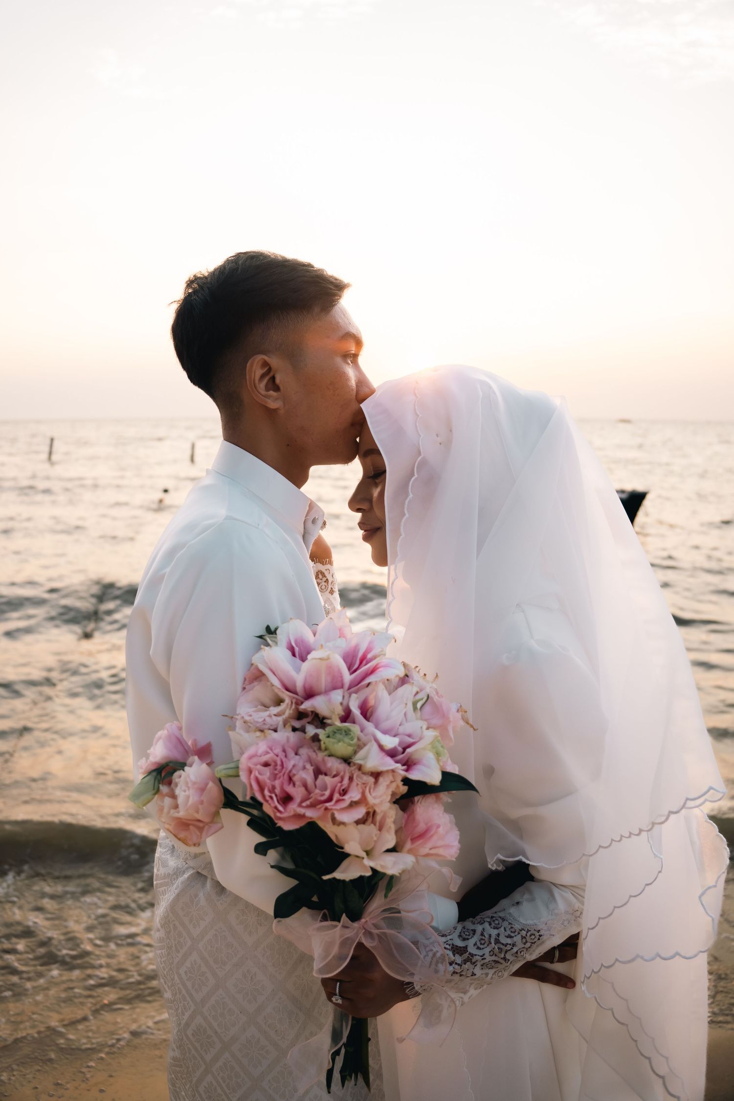 A romantic seaside wedding photo with flowing veil and pink floral bouquet during sunset at the beach.