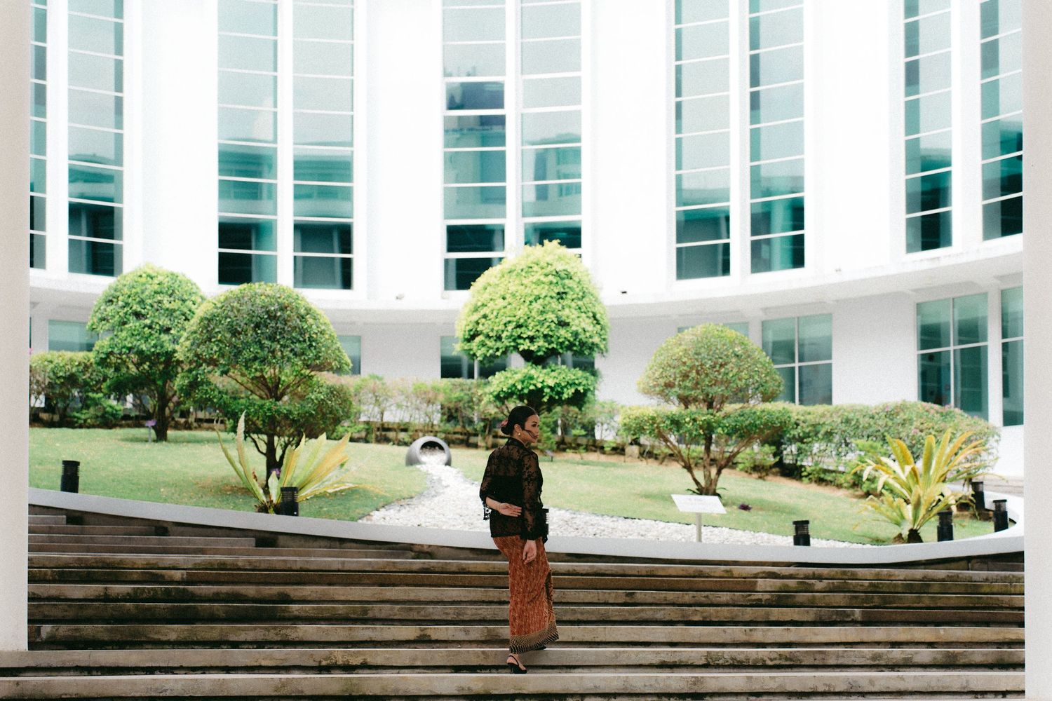 A person stands on concrete steps in front of a modern courtyard with manicured trees and glass windows.
