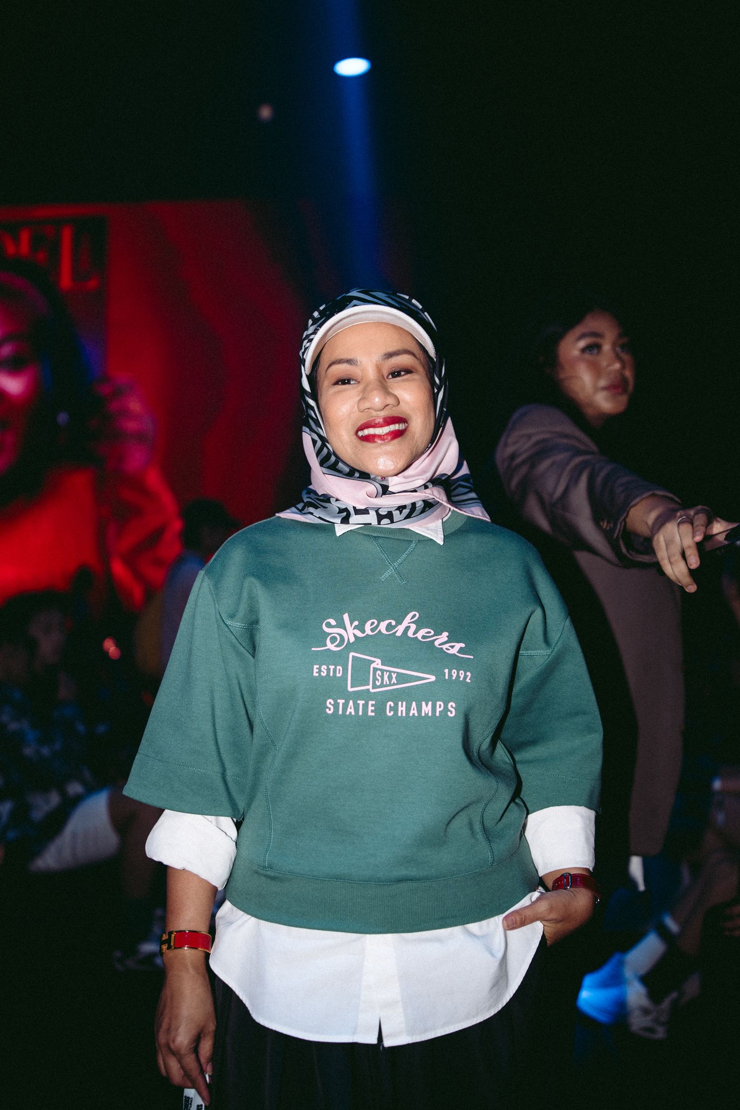 A person wearing a green Skechers sweatshirt and hijab smiles at a fashion event.
