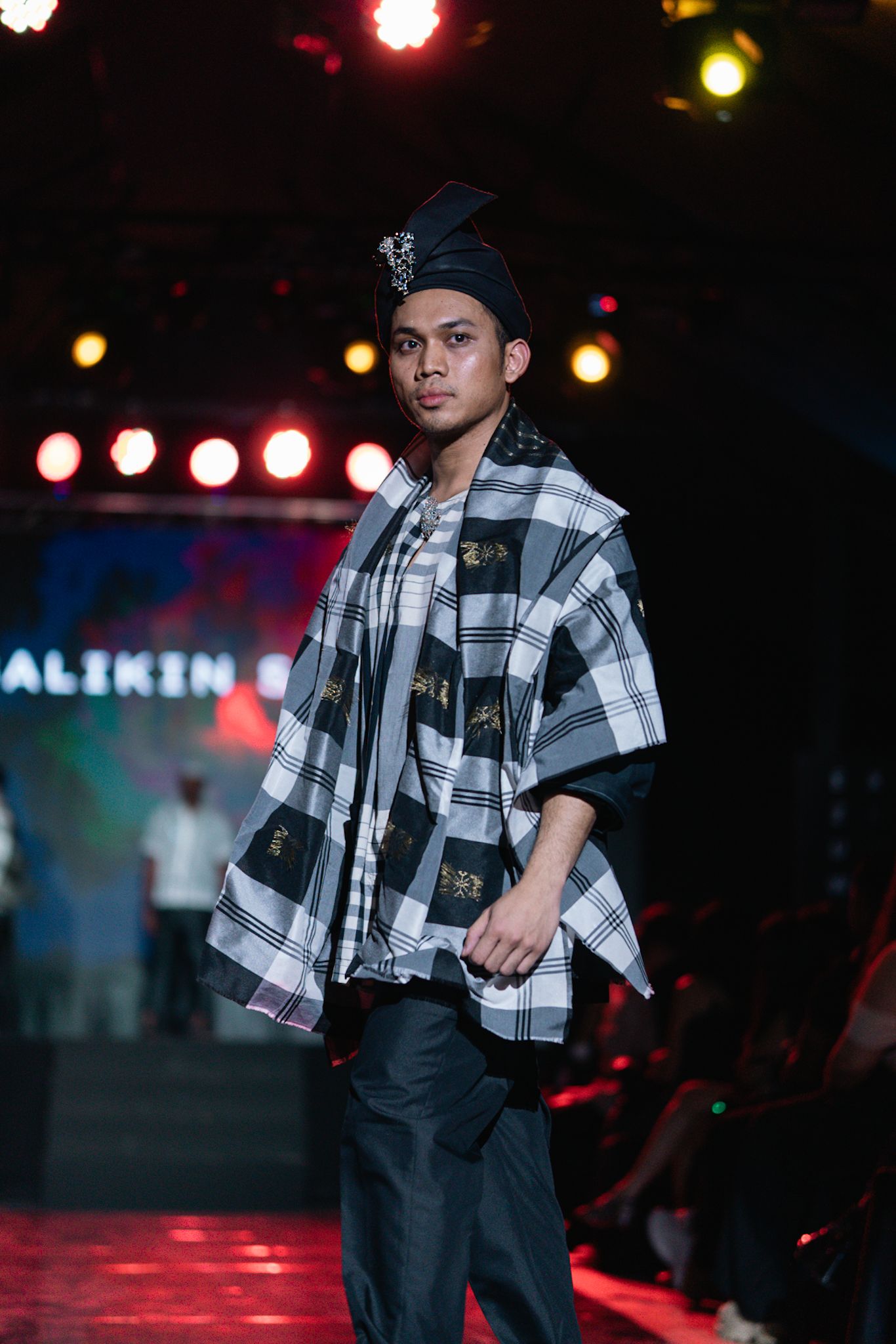 Fashion model wearing plaid poncho and dark pants on runway with colorful stage lighting.