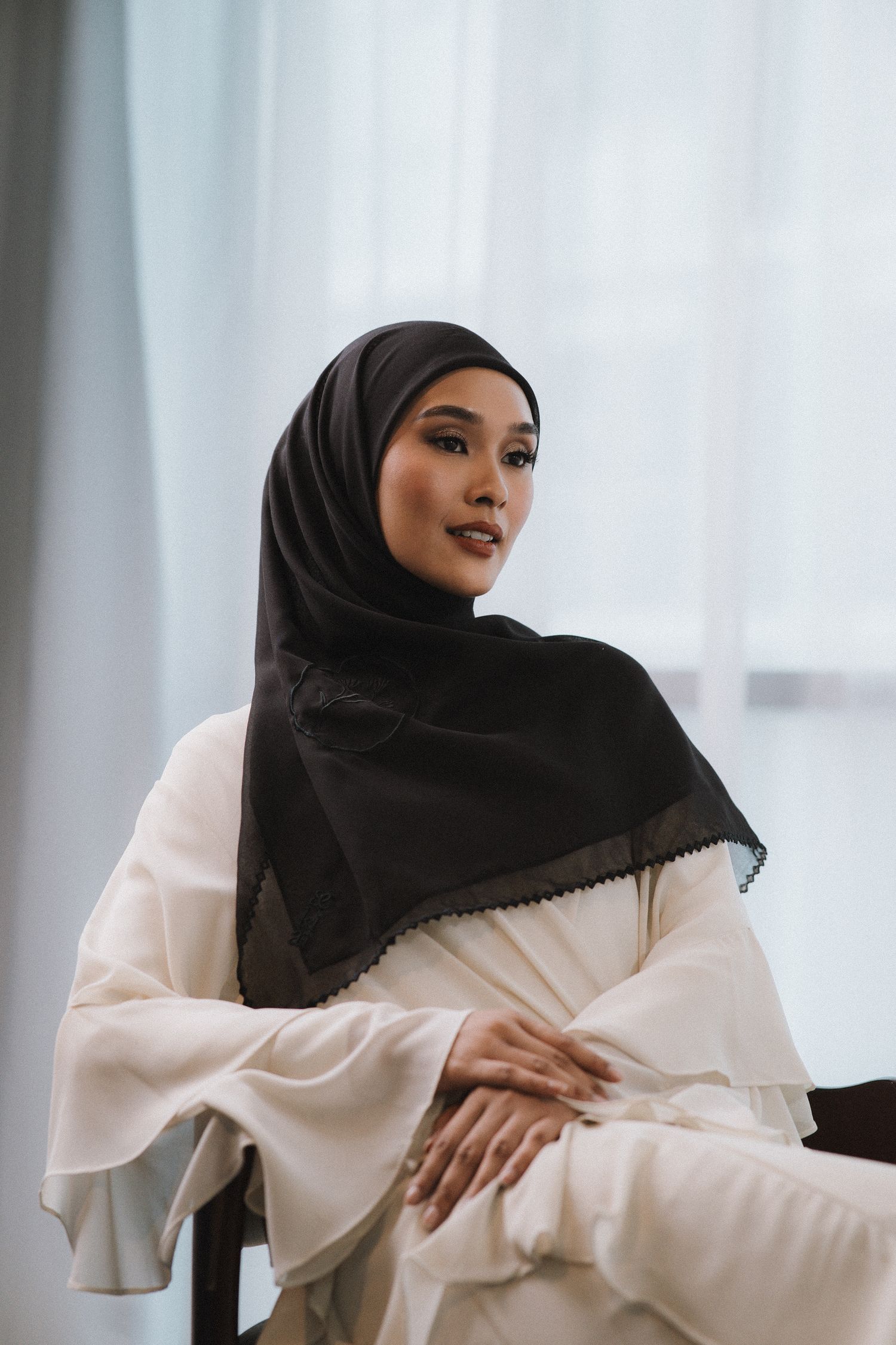Fashion portrait in black hijab and flowing white dress against white curtain backdrop.