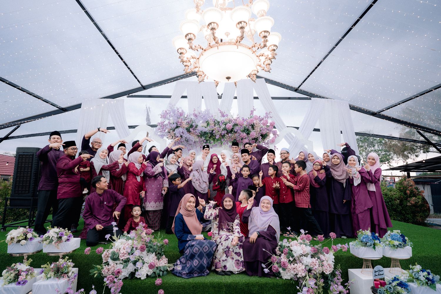 Large group in matching purple outfits posing under chandelier at floral-decorated outdoor wedding celebration.