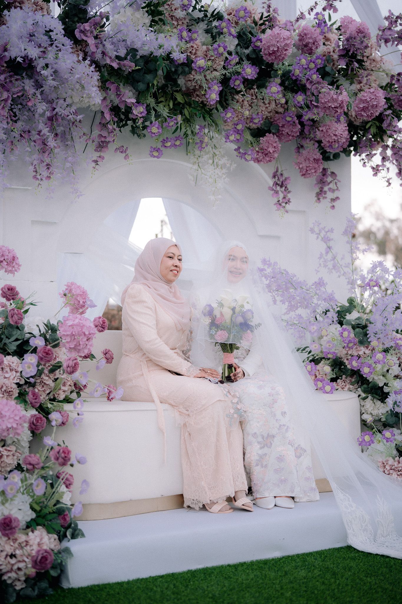 Two women in hijabs pose at a floral wedding arch adorned with purple and white flowers.