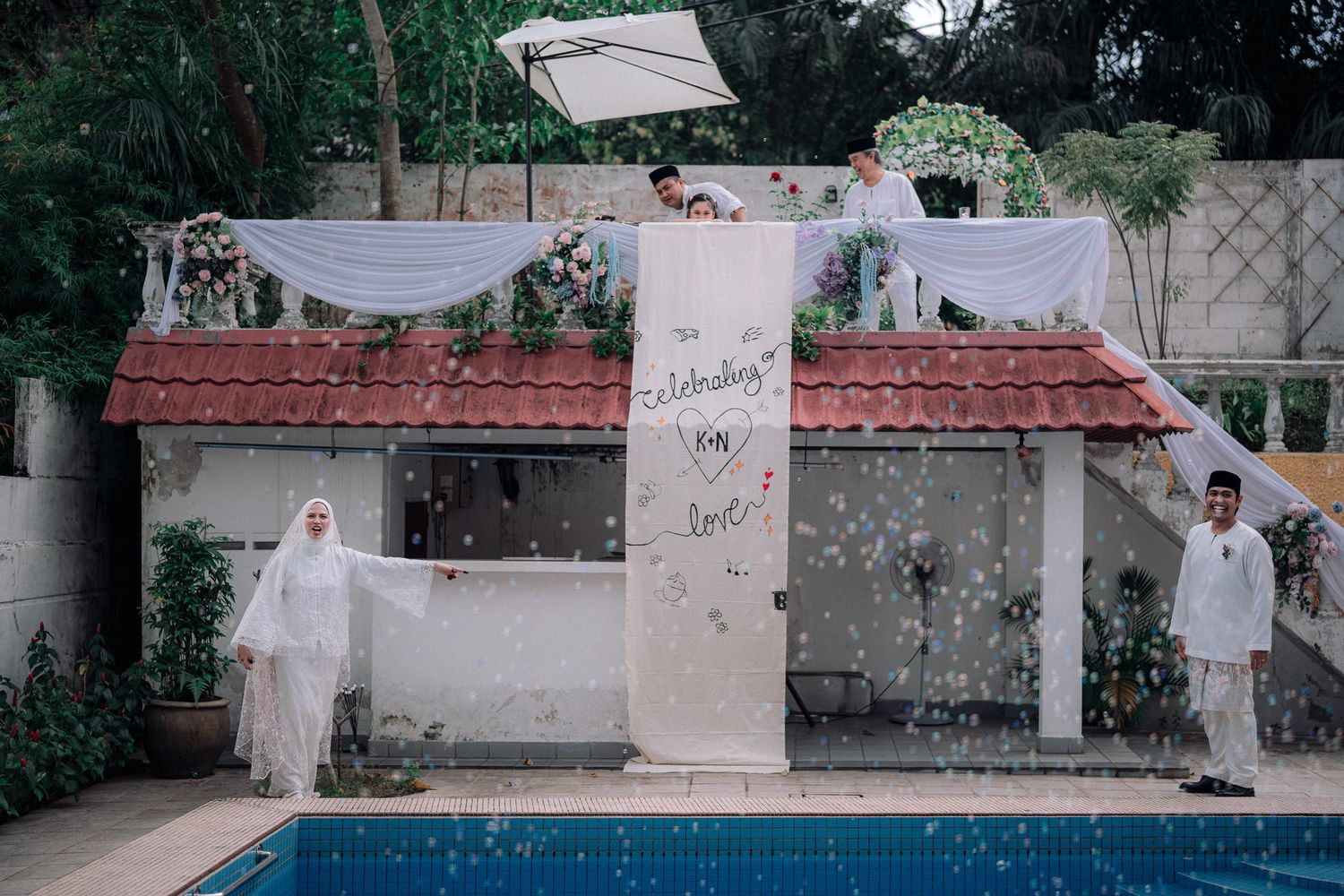 Muslim wedding couple in white attire celebrating by a pool with floral decorations and a celebratory sign.