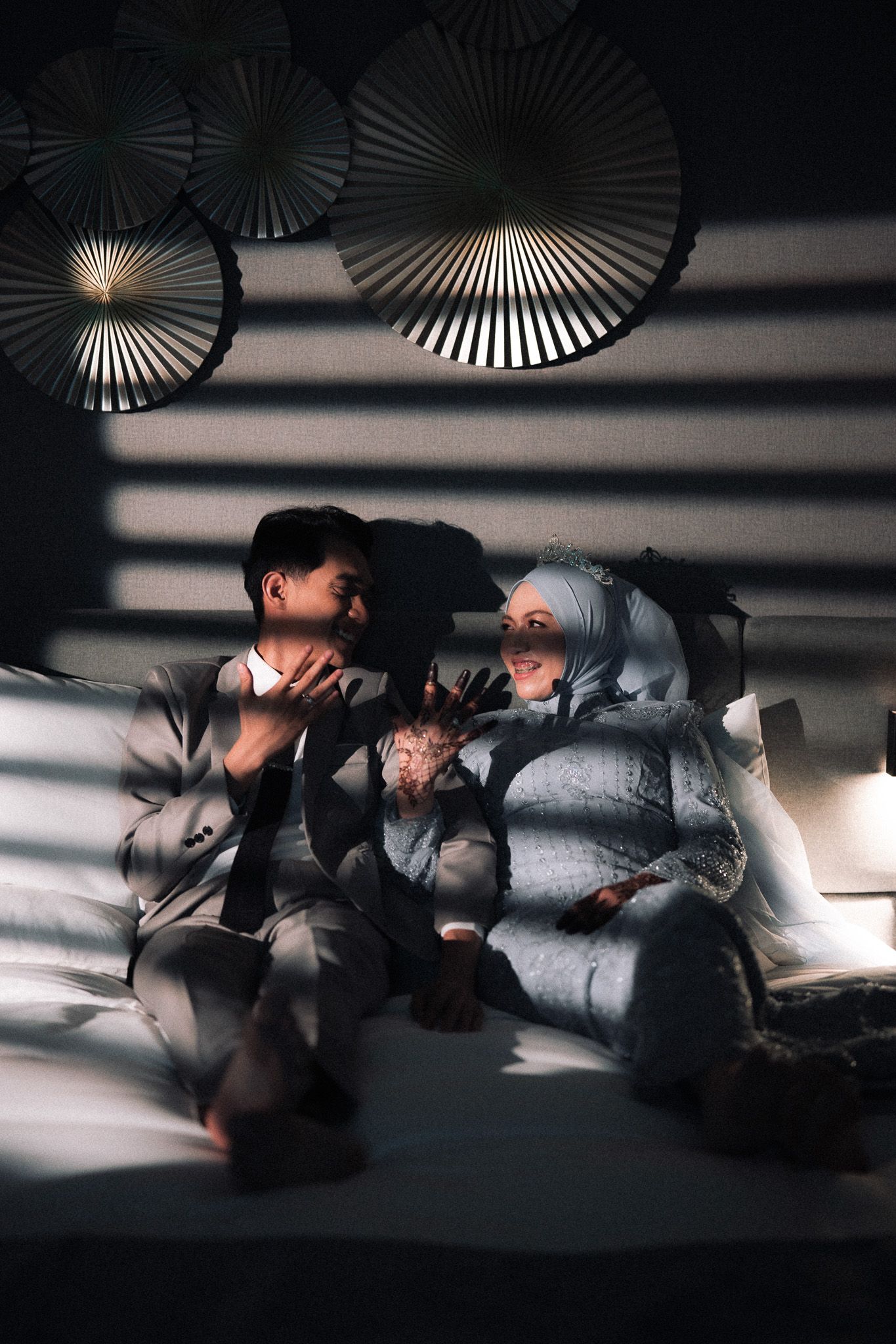 Muslim couple sitting on a bed in dramatic light and shadow, smiling and talking, with decorative paper fans on the wall.