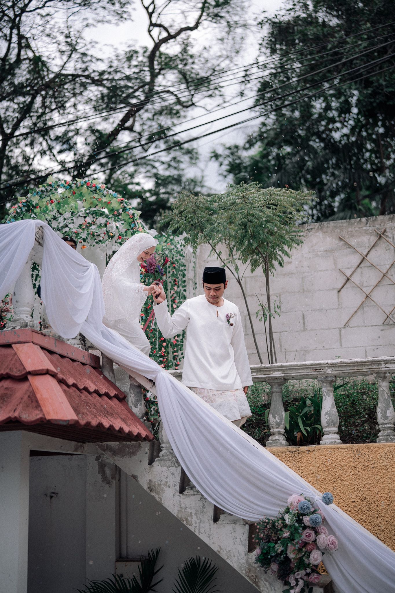Muslim bride and groom in white attire descending decorated stairs with floral arrangements at outdoor wedding.