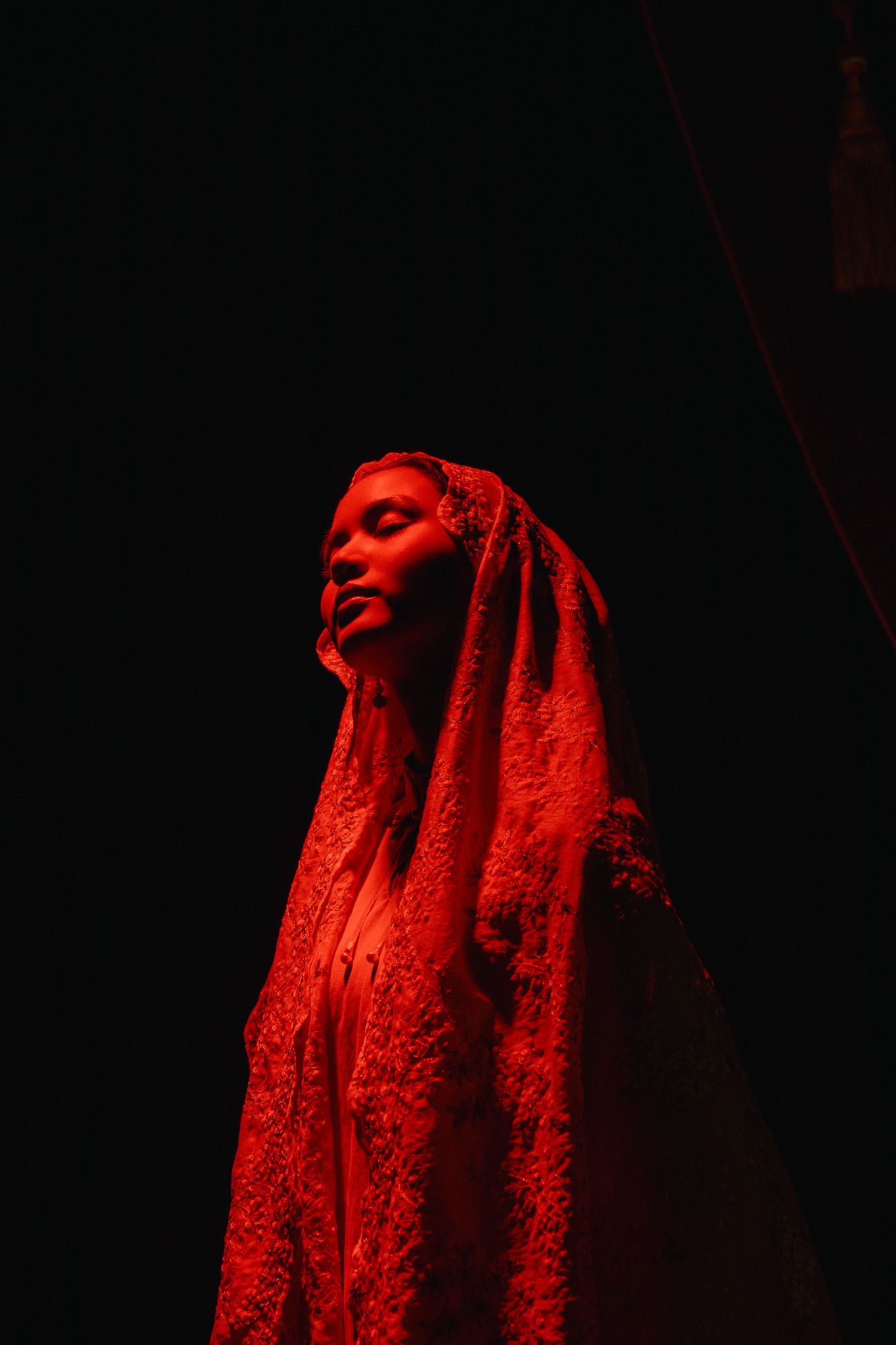 Person draped in white lace veil illuminated by dramatic red lighting against dark background.