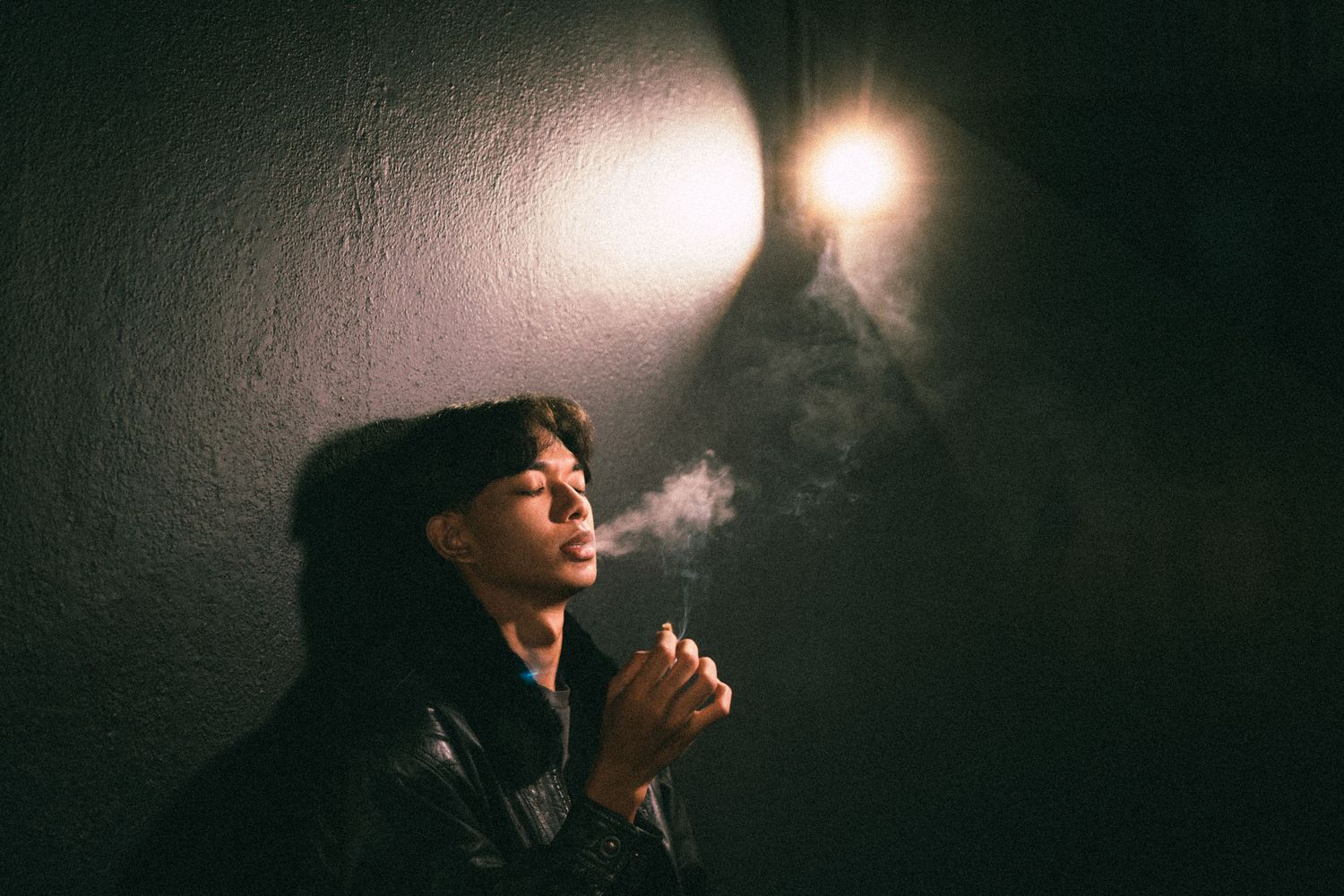 Atmospheric portrait of someone smoking under dramatic lighting against a dark wall.