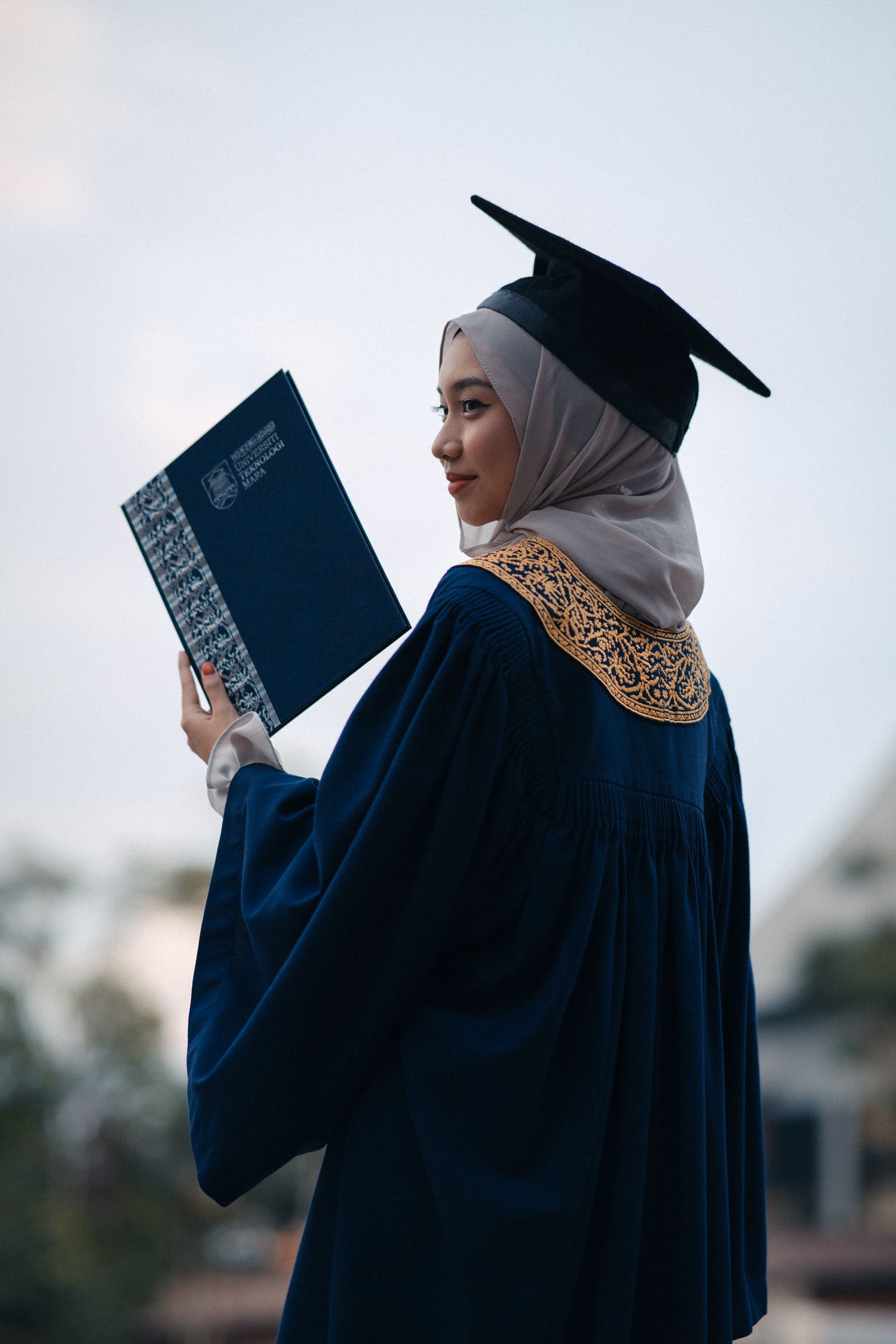 Graduate in blue gown with gold embroidery holding diploma against bright sky.