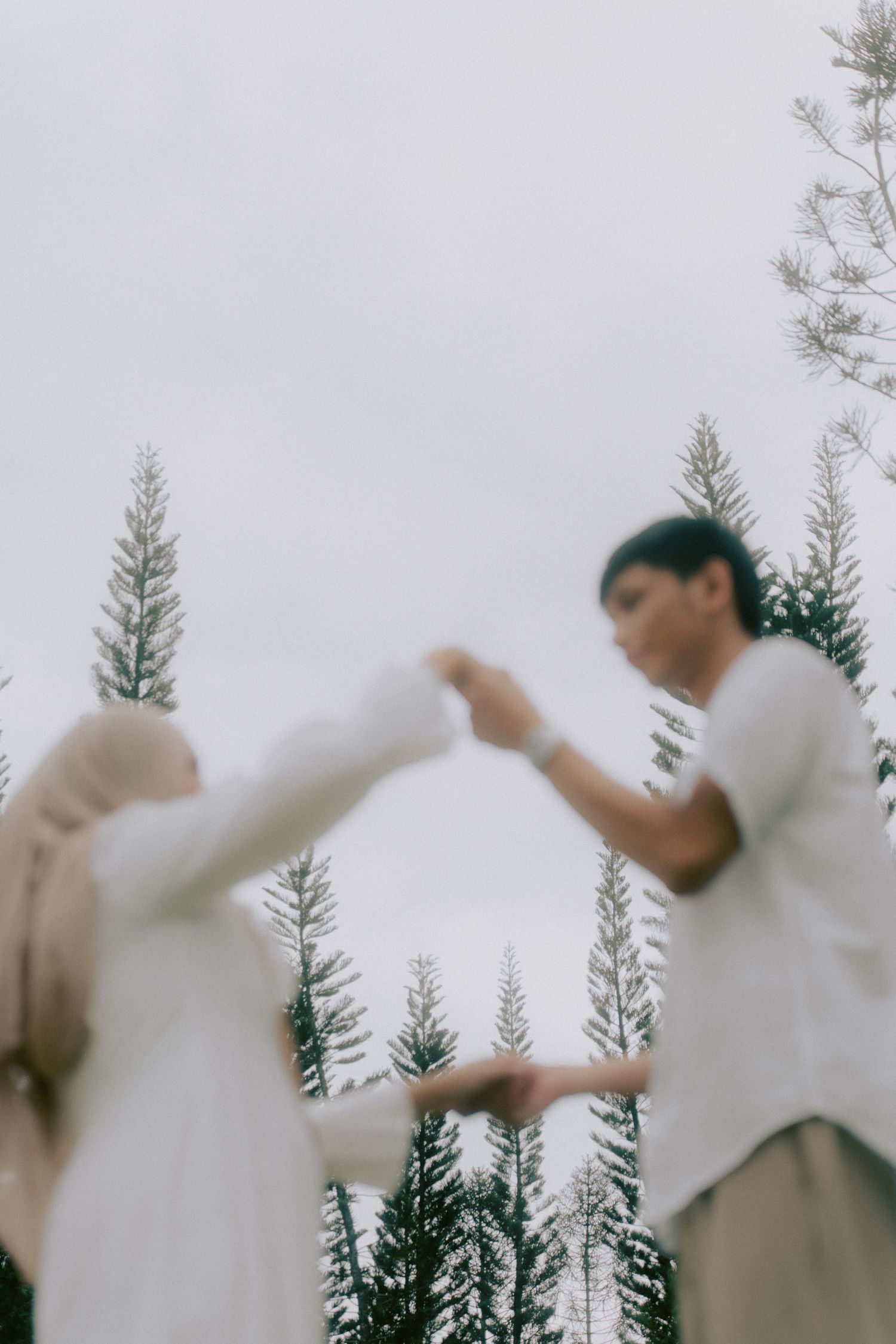 Two people in white outfits dance together against a backdrop of evergreen trees on an overcast day.