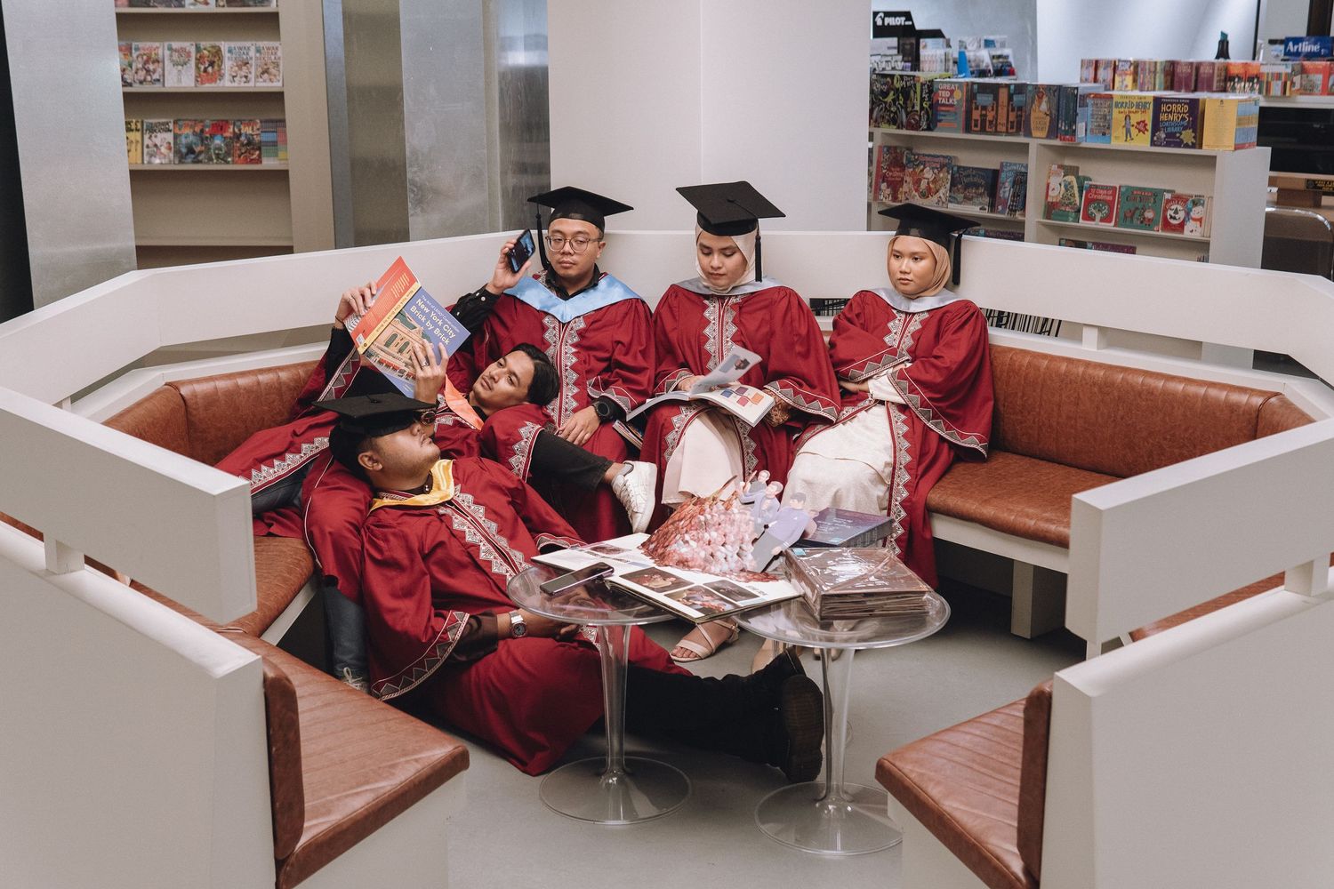 Graduates in red robes relax and read together on circular seating in a library.