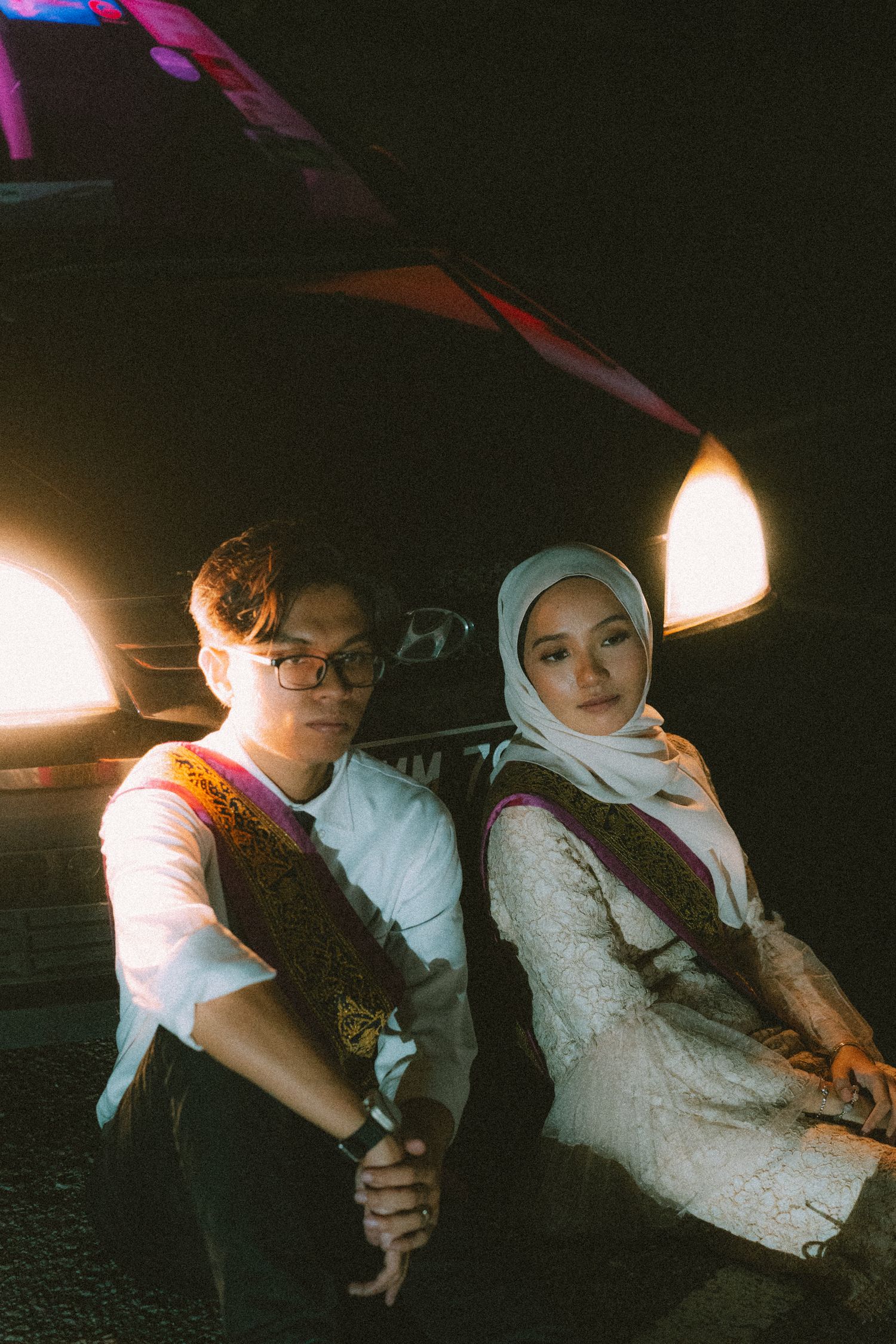 A couple poses together near car headlights at night.