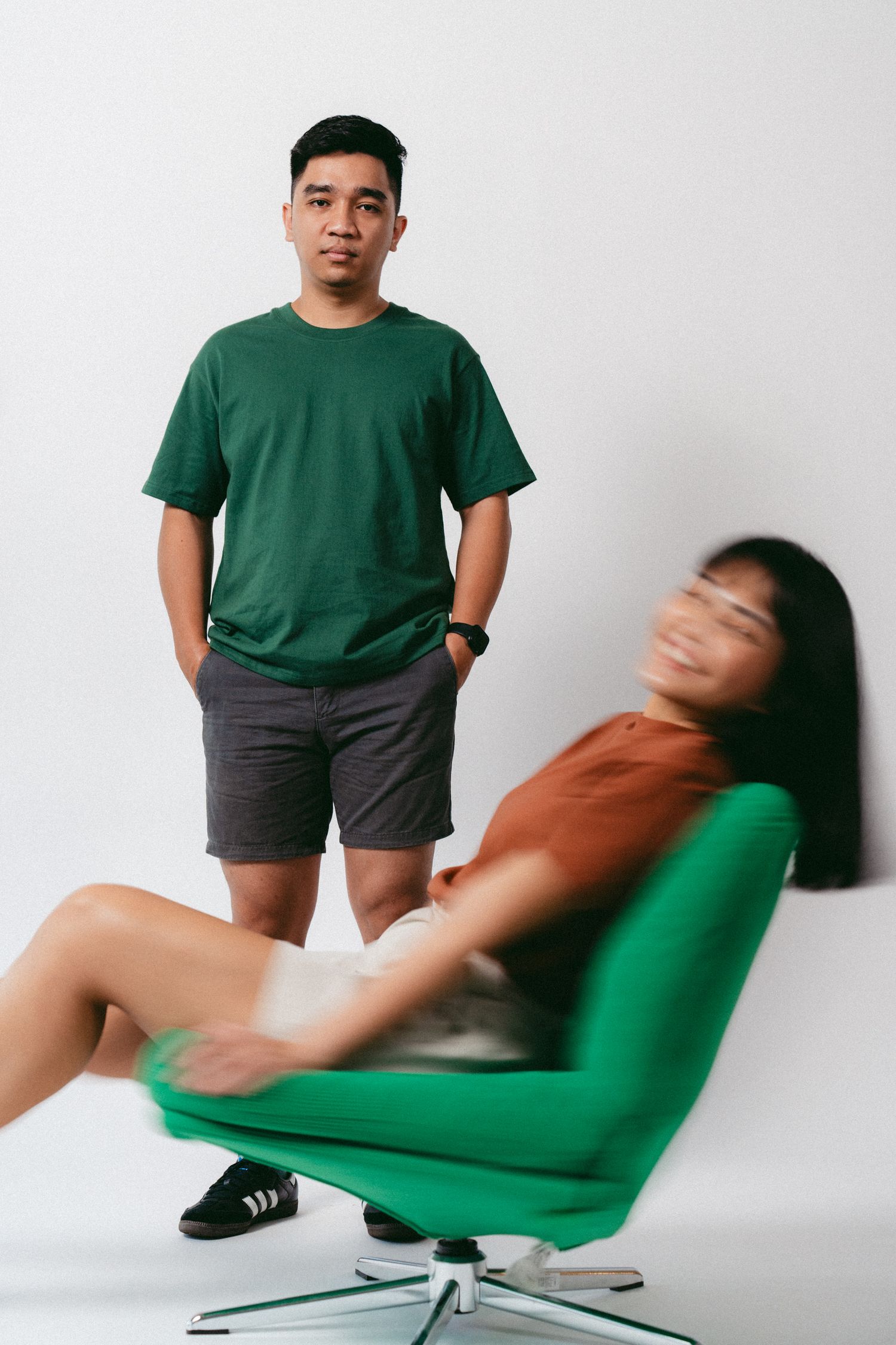 Casual couple posing with person spinning in green office chair against white background.