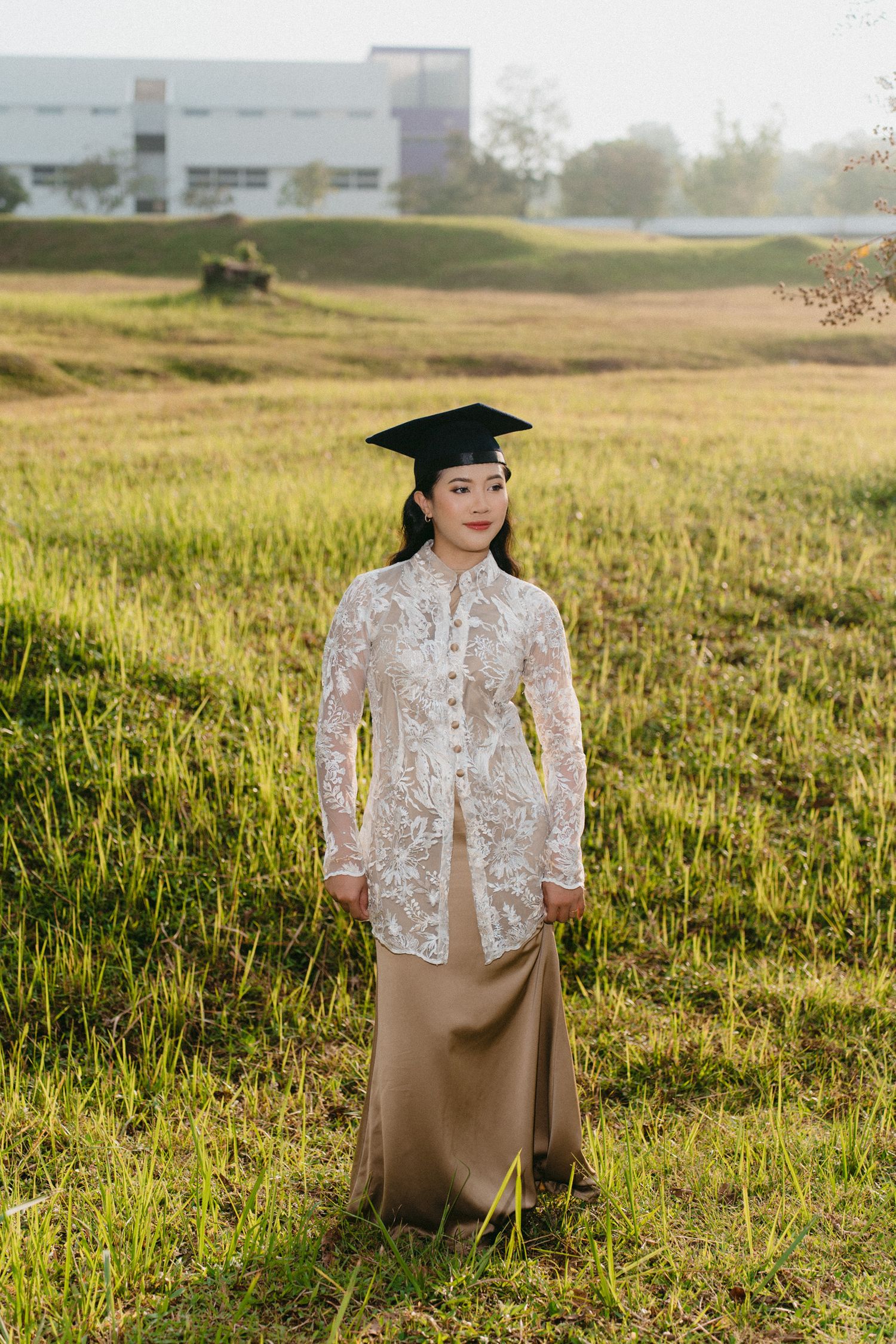 Graduate in a white lace jacket and beige dress poses in a sunlit grassy field during golden hour.