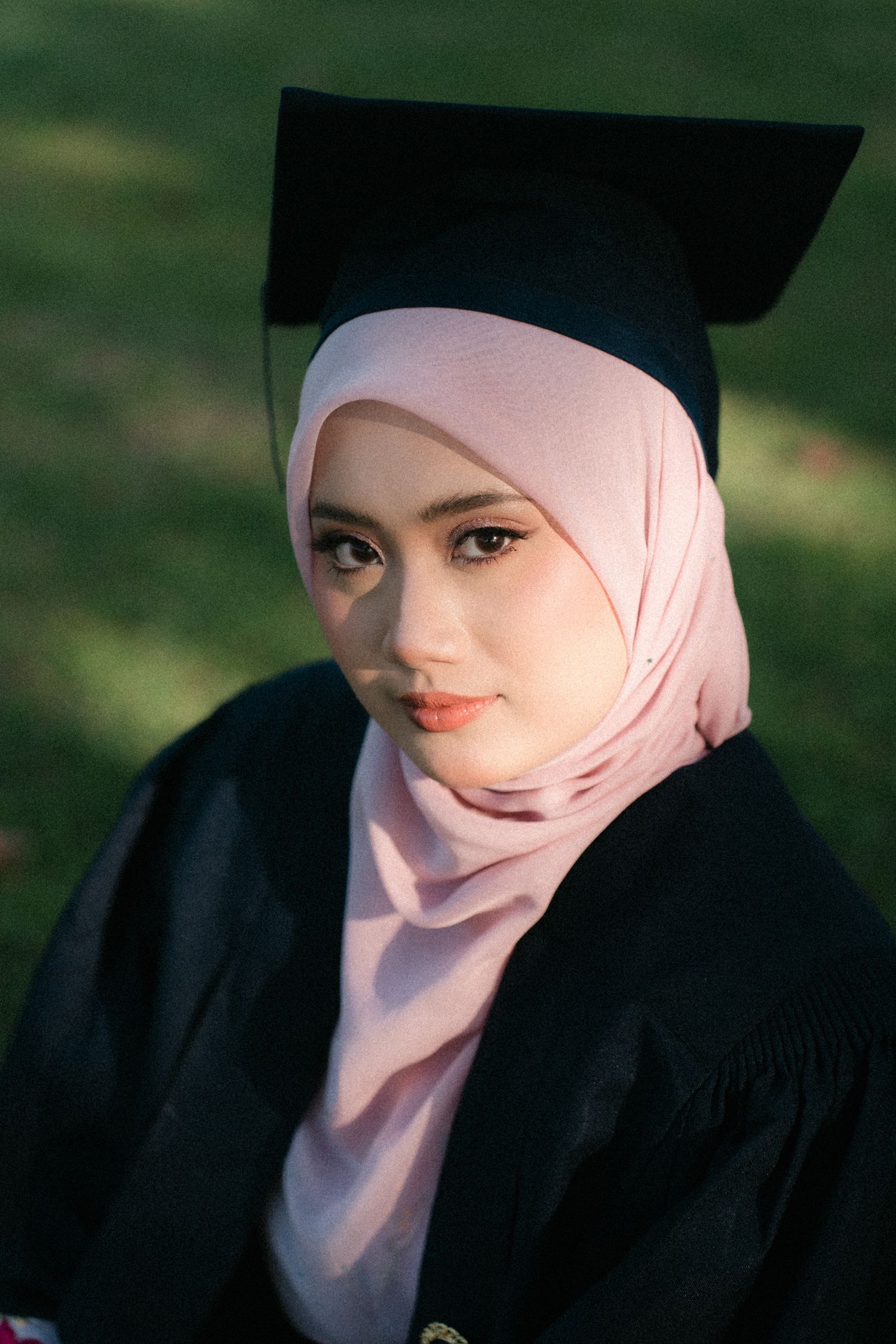 Graduate in black cap and gown wearing a light pink hijab poses for a portrait against a blurred green background.