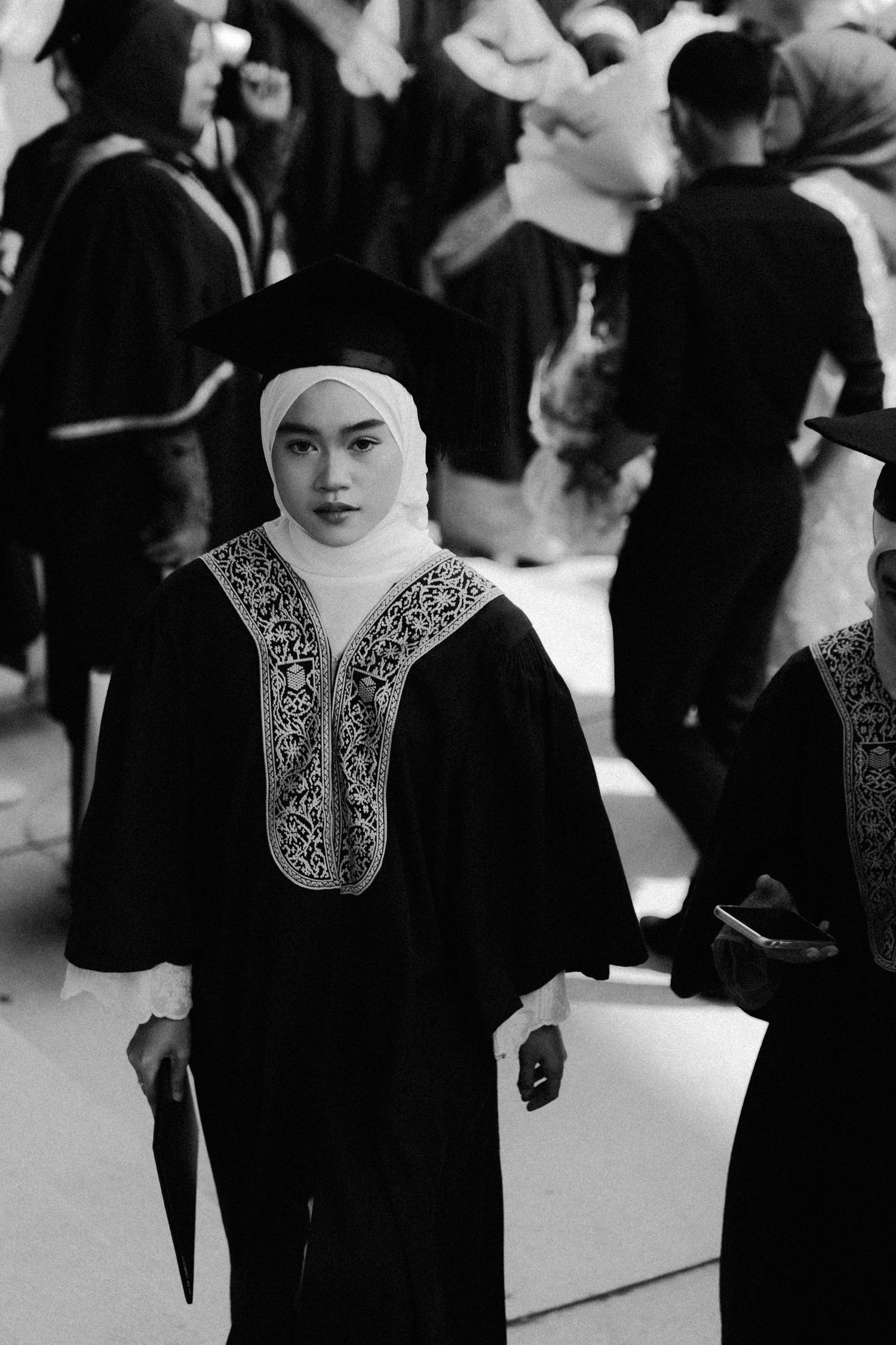 A figure in traditional Middle Eastern dress with embroidered details stands amid a crowd in black and white photography.