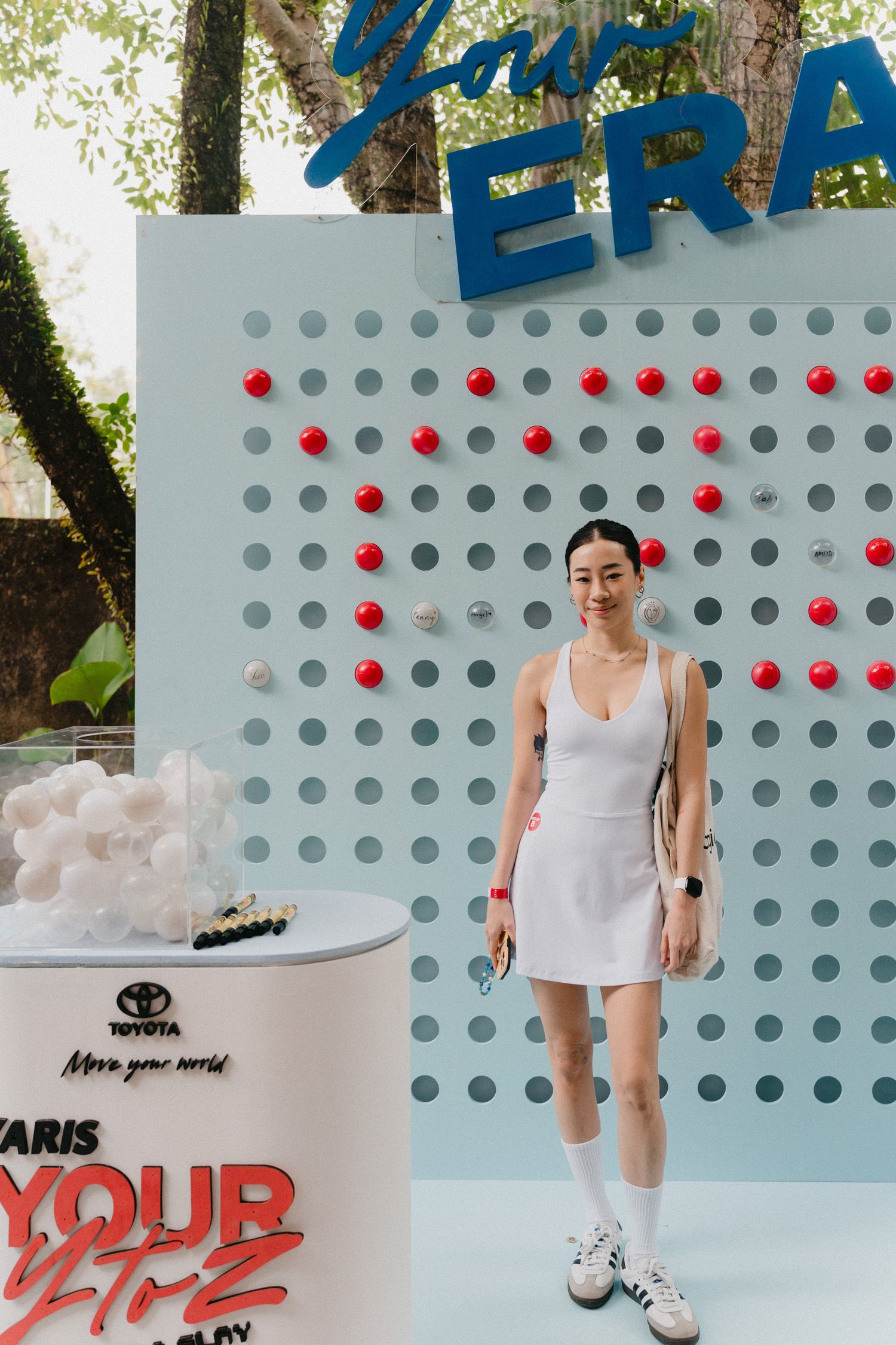 A person in white clothing stands against a polka dot wall with red dots in an outdoor setting.