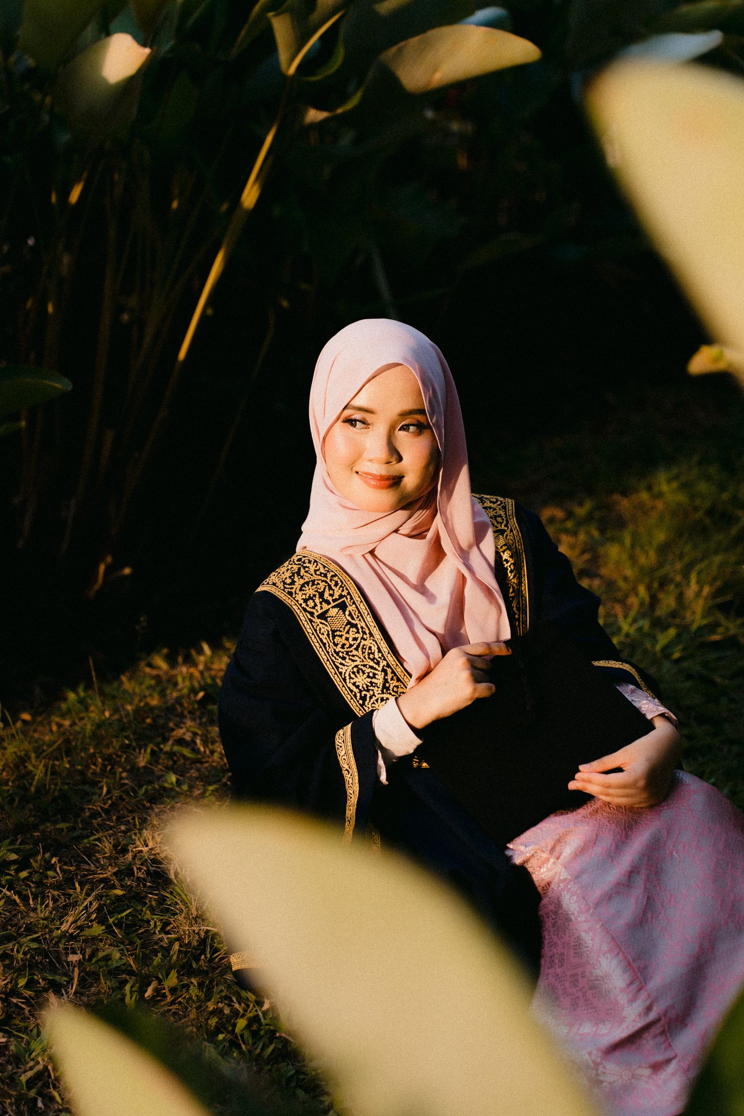 A person in a pink hijab and purple outfit poses thoughtfully in a natural outdoor setting with dramatic lighting.