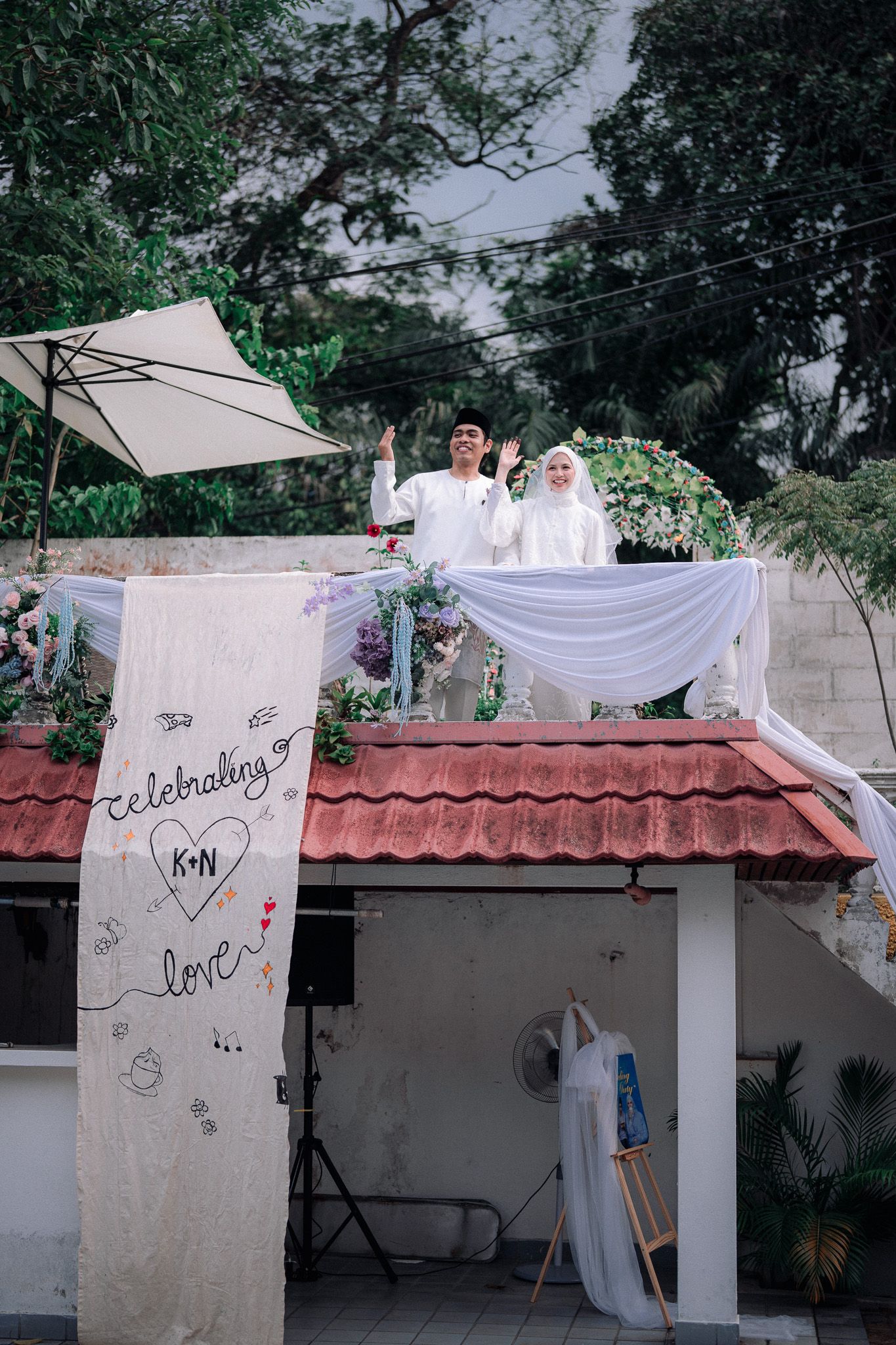 Muslim couple in wedding attire waving from rooftop decorated with flowers and white drapes, celebrating love outdoors.