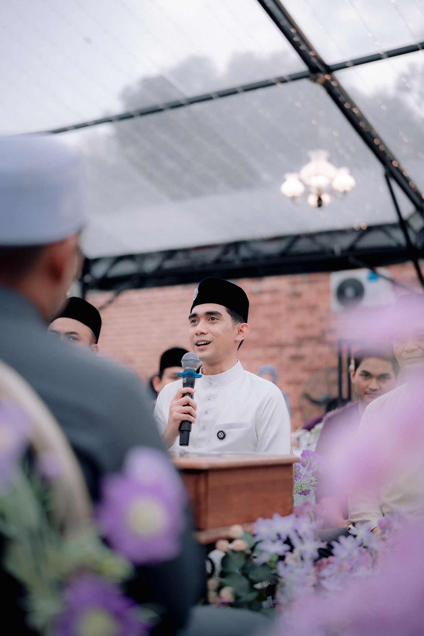 Young man in white baju melayu and songkok speaks at a Malay wedding ceremony with purple floral decorations.