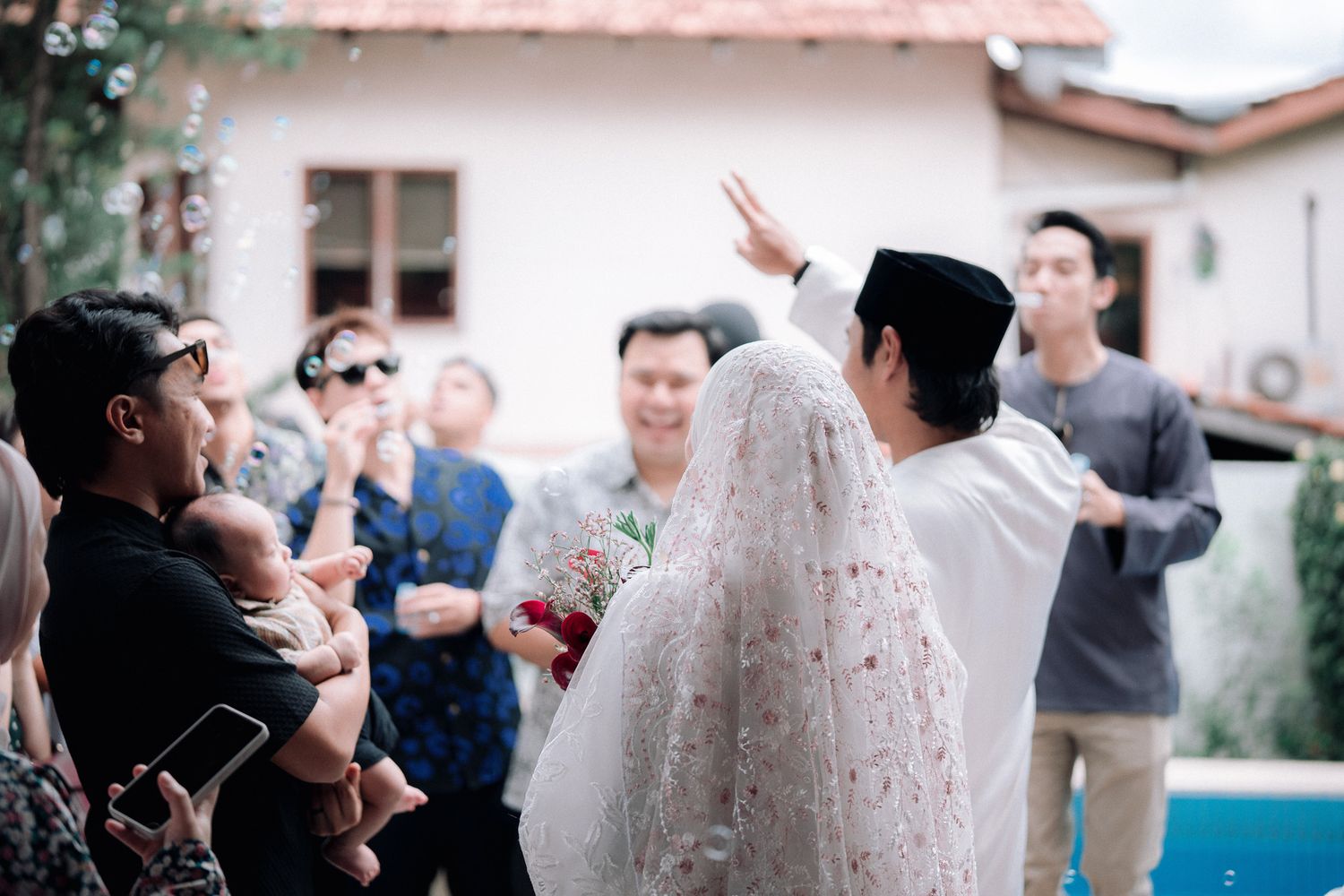 A couple in traditional Muslim wedding attire celebrates outdoors with guests, bubbles floating in the air near a pool.