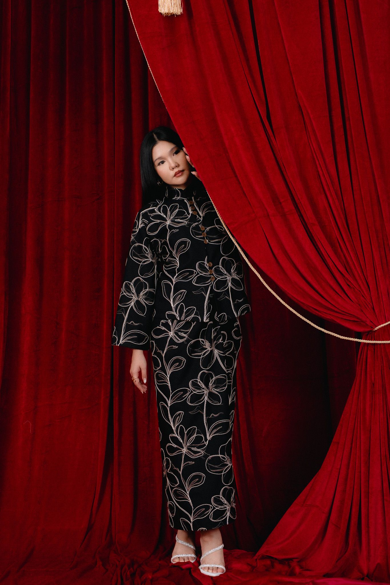 Woman in black floral embroidered cheongsam dress standing between red velvet curtains.
