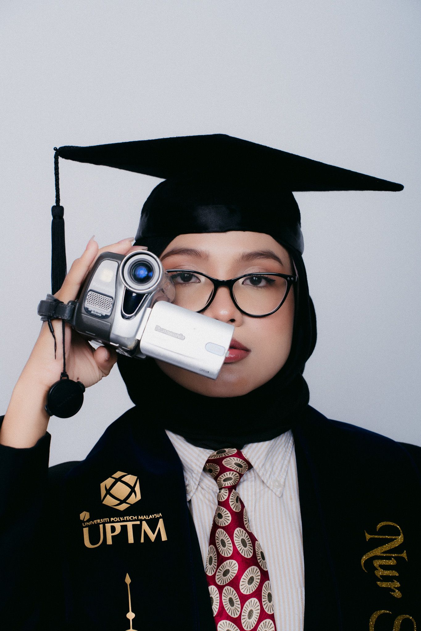 Graduate in UPTM cap and gown holding vintage camcorder, wearing hijab and glasses, posing for graduation photo.