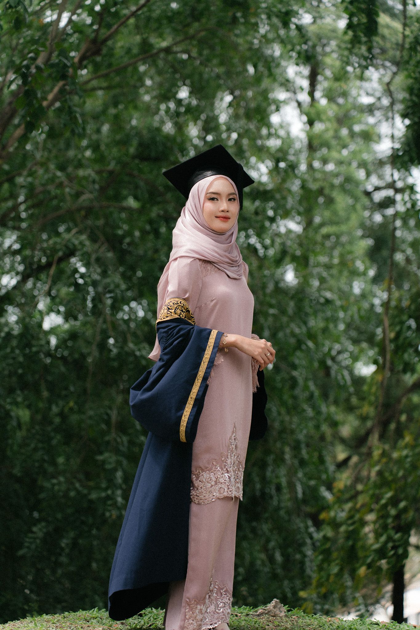 Young Muslim woman in hijab and graduation cap and gown smiling outdoors among green trees.