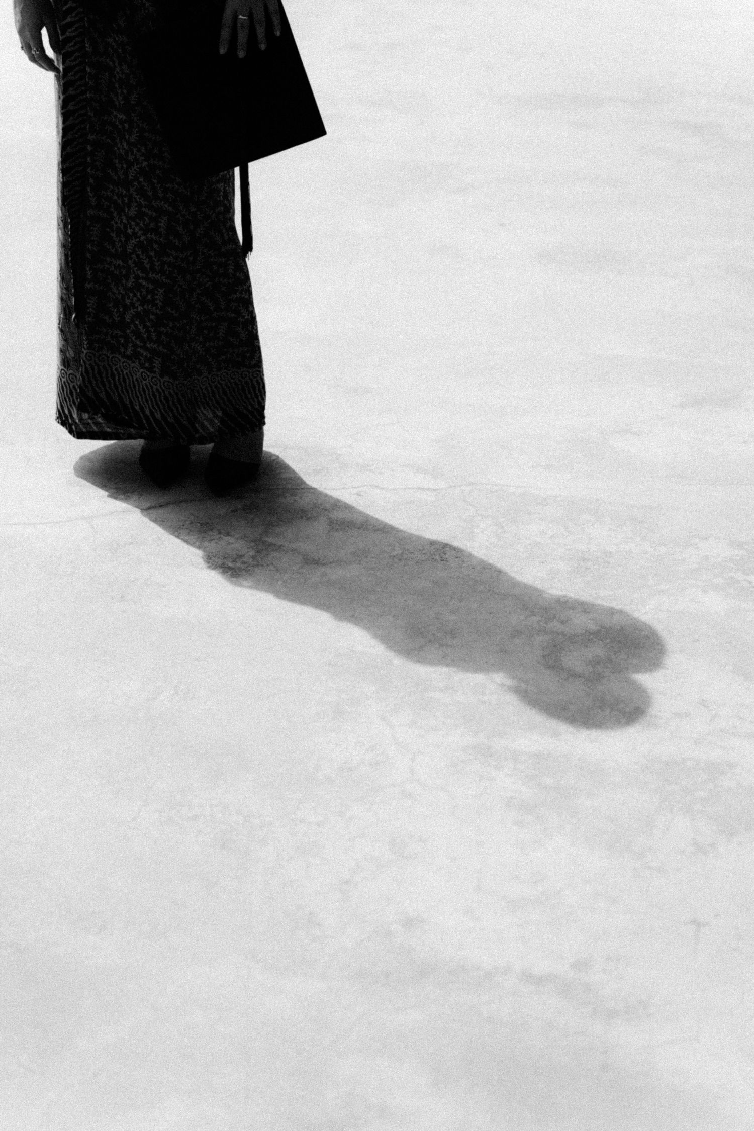 Black and white photograph showing a long dark shadow cast by someone wearing a long dress or robe on a light floor.