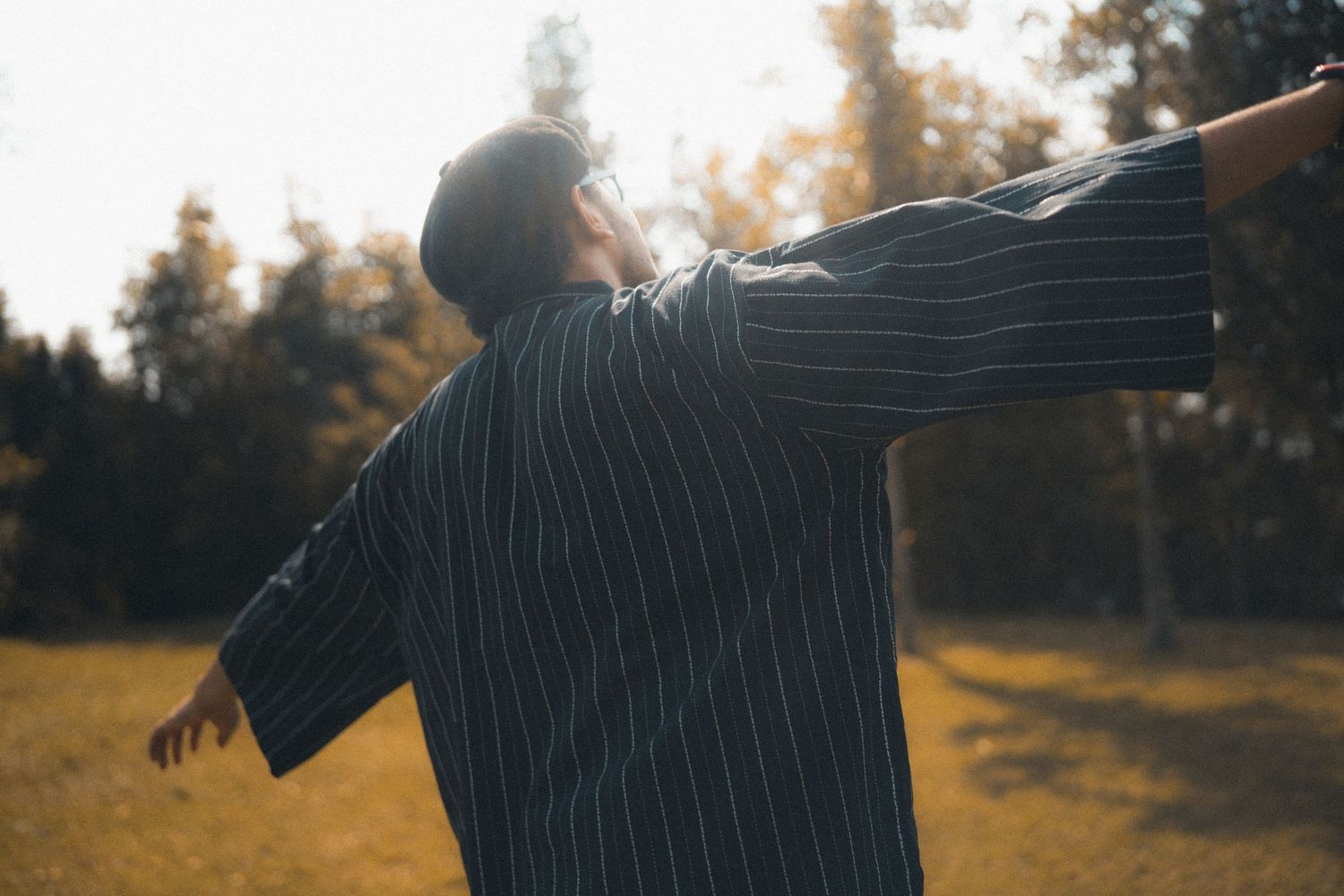 Person in striped shirt stretches arms wide against forest backdrop at sunset.