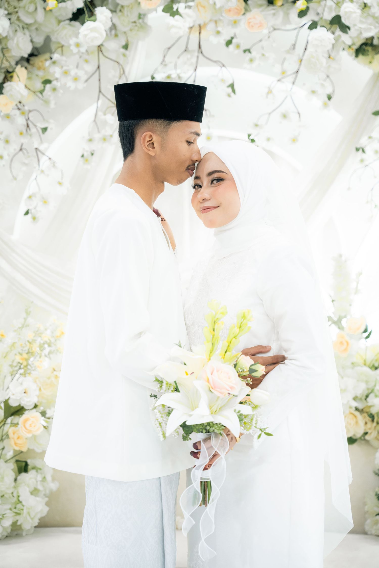 Traditional Malay wedding photo with bride in white hijab and groom in baju melayu against floral backdrop.