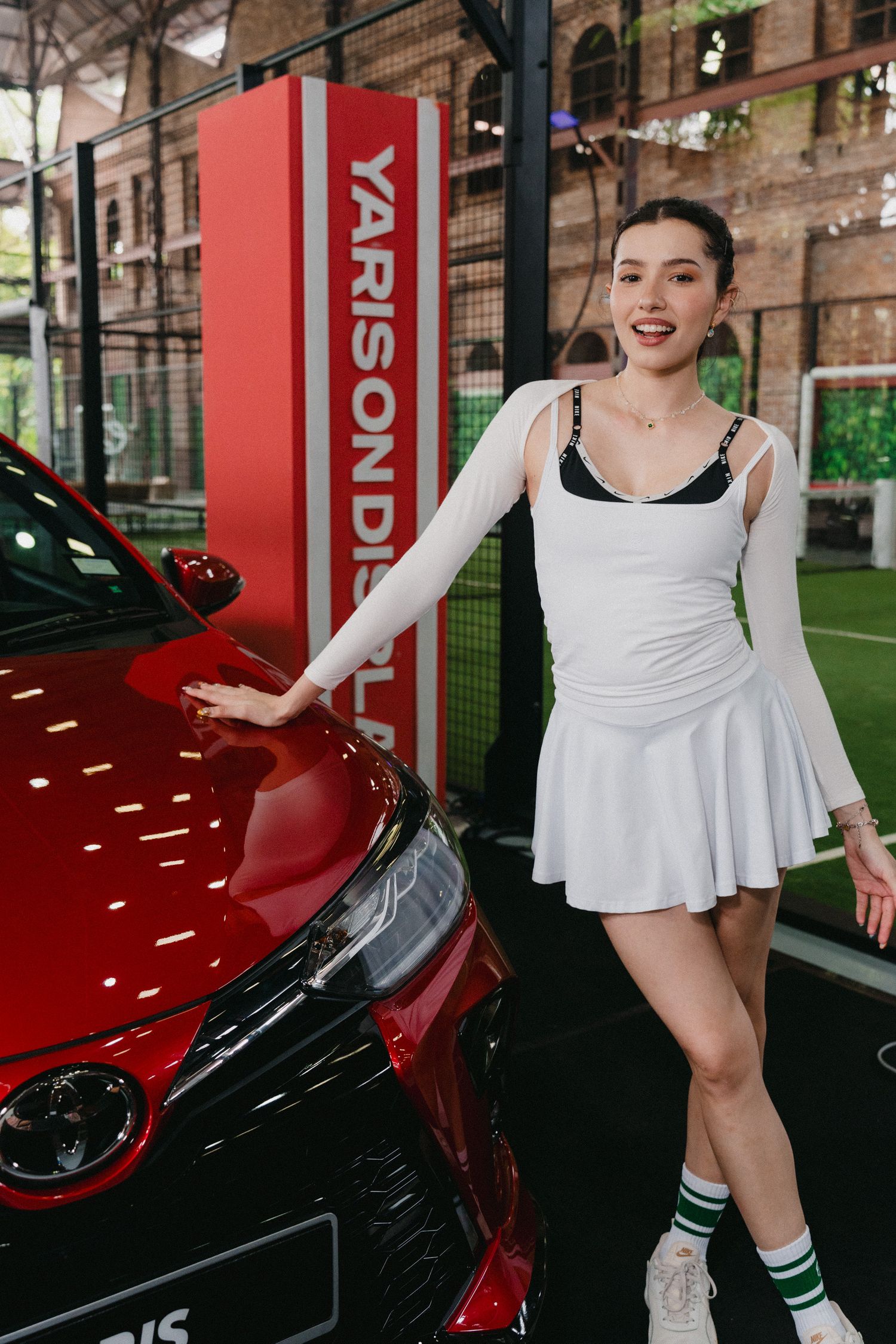 A person in a white tennis dress poses playfully next to a red sports car in an indoor setting.