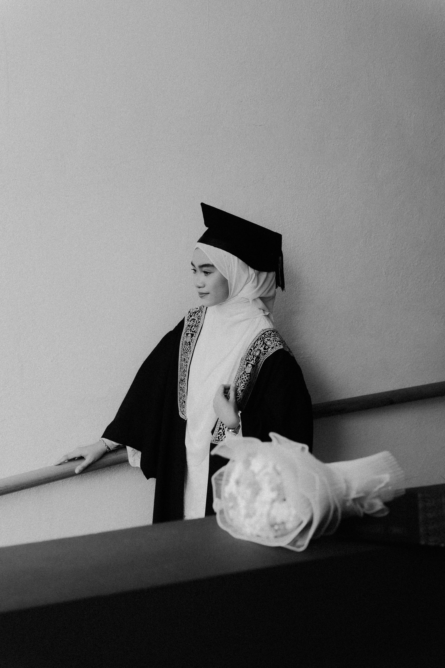 A graduate in academic regalia stands in a hallway holding a wrapped gift package in black and white photography.