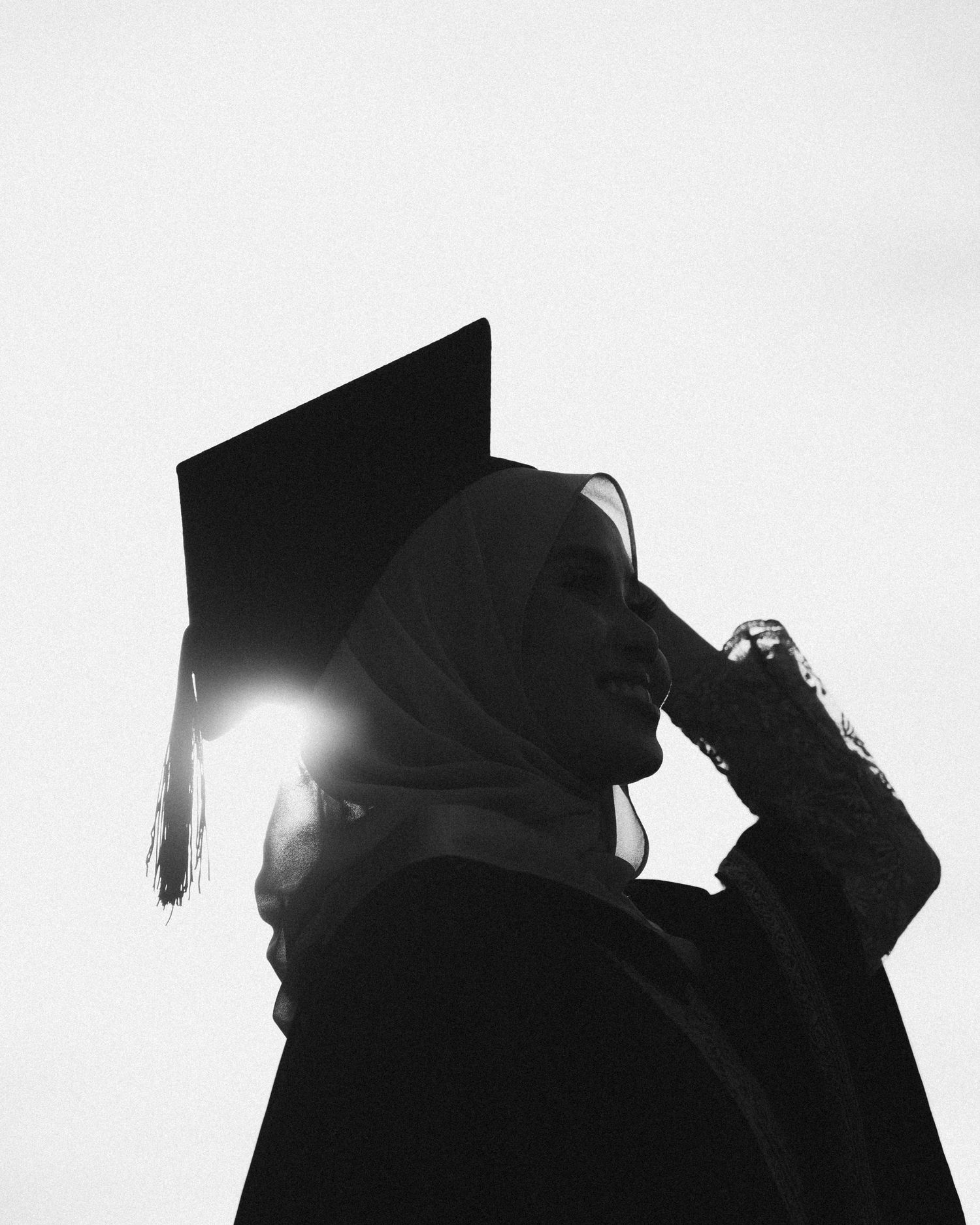 Black and white silhouette of a graduate in cap and gown against bright sunlight creating a dramatic backlit effect.