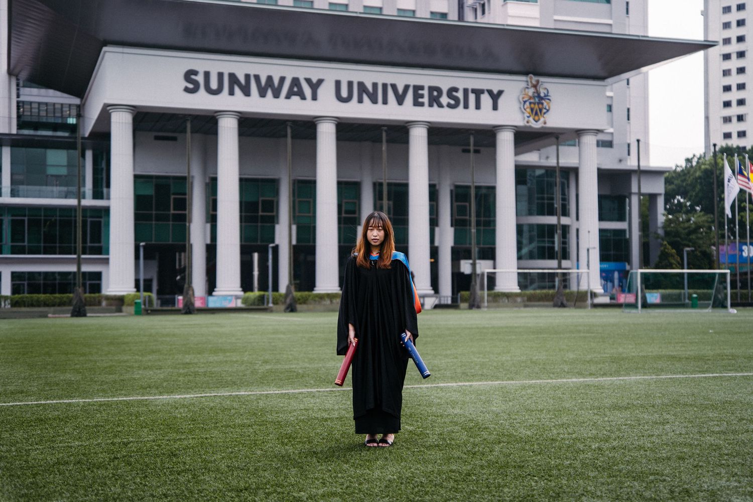 Graduate in traditional academic regalia standing alone on green lawn in front of Sunway University.