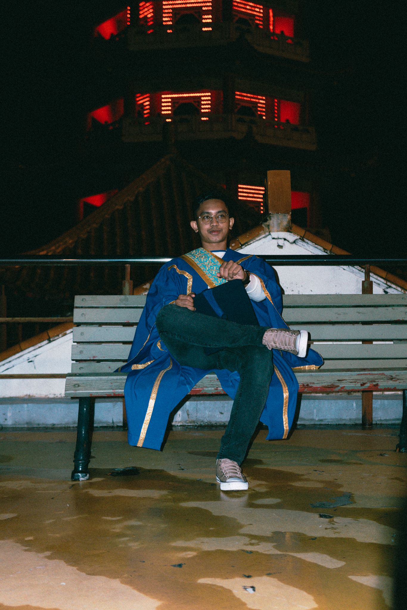 Graduate in blue regalia seated on bleachers with illuminated pagoda tower in background.