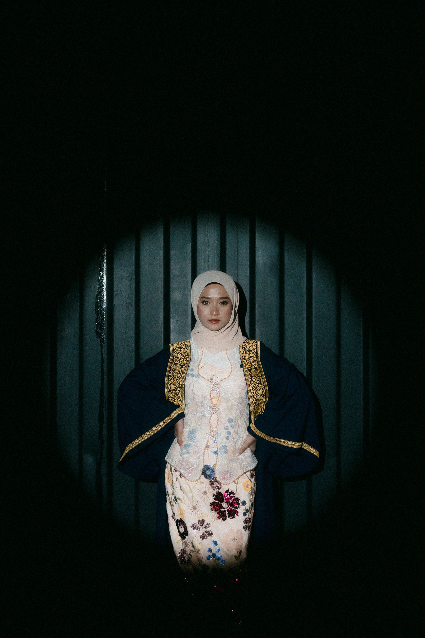 A Muslim woman in a white hijab and floral dress with a dark blue gold-trimmed robe stands in a circular spotlight.