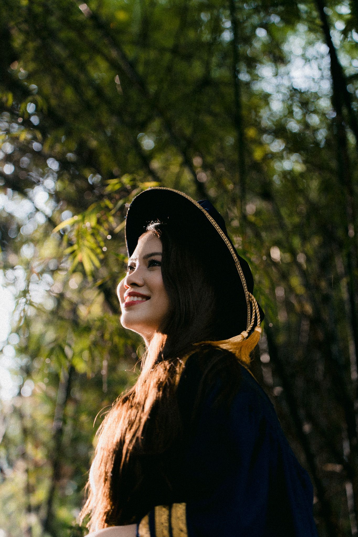 Smiling young woman in black graduation gown and cap stands outdoors in sunlit bamboo forest.