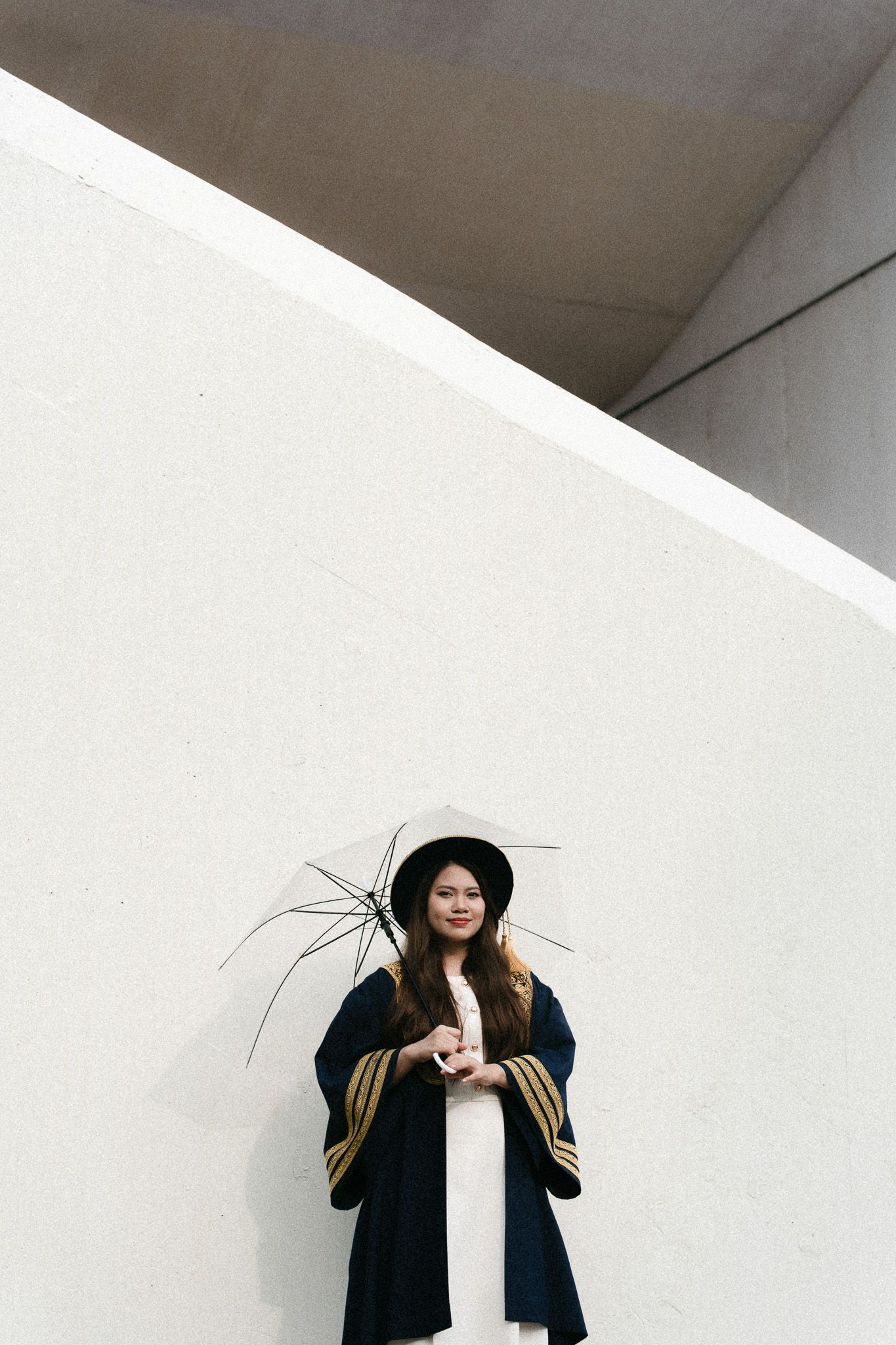 Young Asian woman in navy graduation robe and hat holds umbrella against white wall.