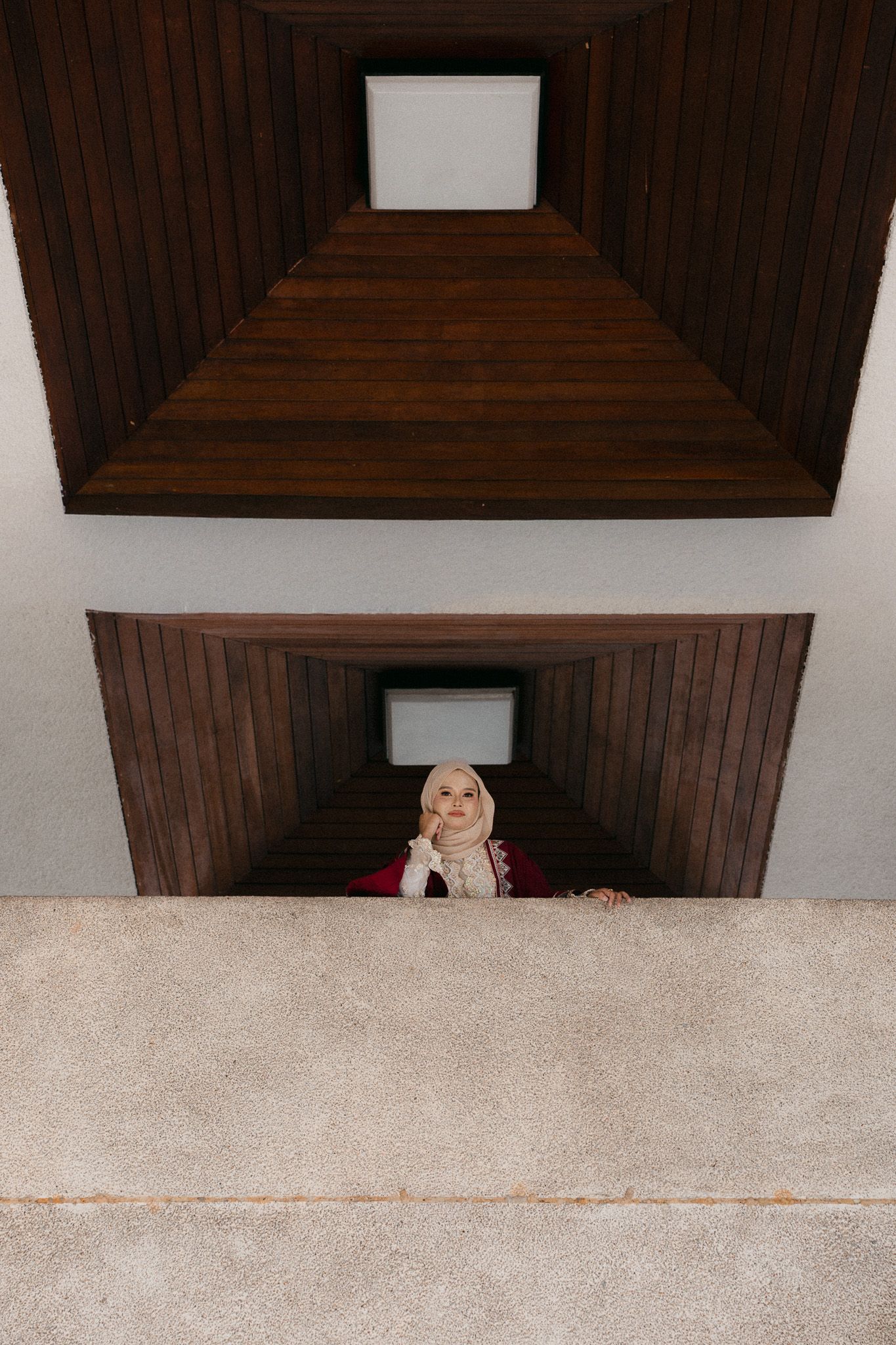 Woman in hijab looking up through geometric wooden stairwell architecture with symmetrical perspective.