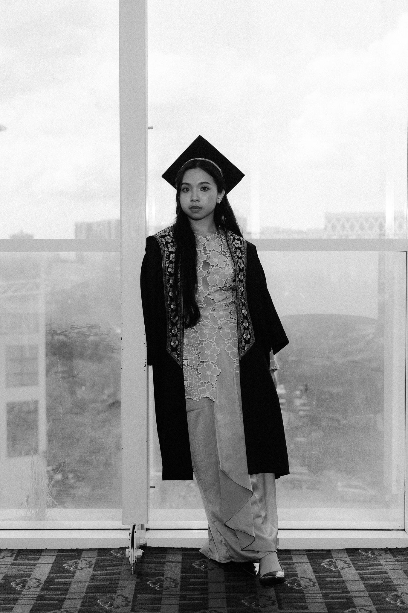 Black and white photo of a young woman in graduation gown and mortarboard cap standing by large windows.