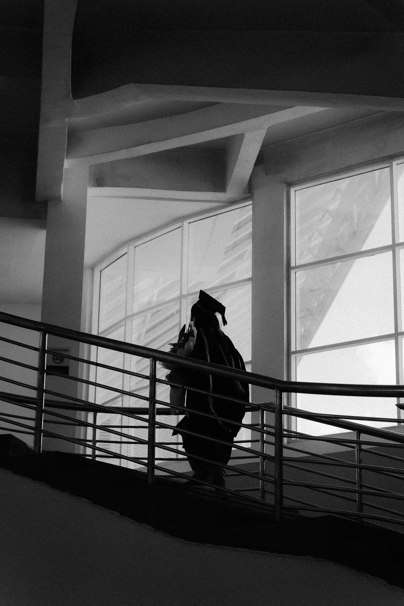 Black and white photo of a graduate in cap and gown silhouetted on a staircase near large windows.