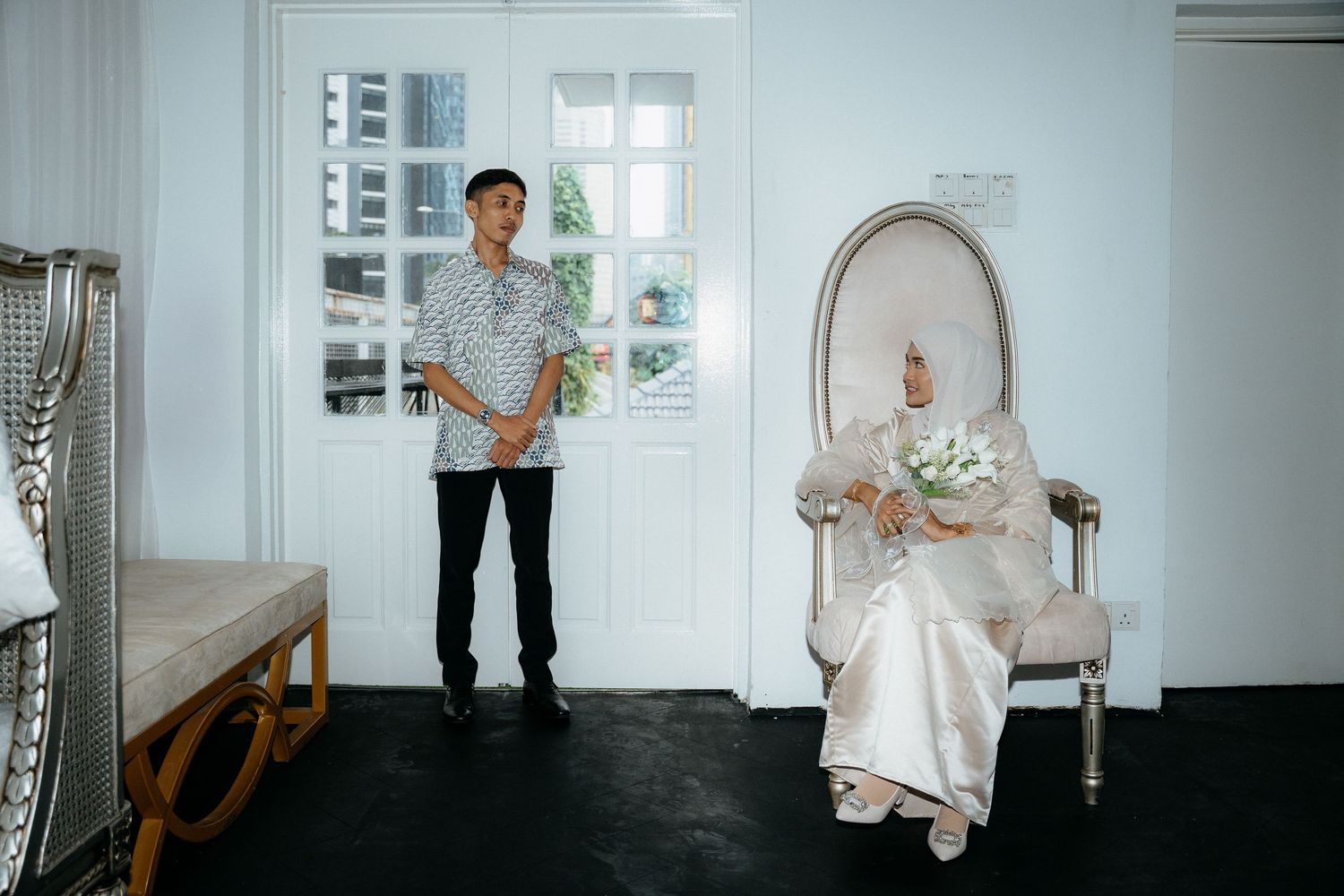 Two figures in a bright white room, one standing near a window and another seated in an ornate chair with floral decor.