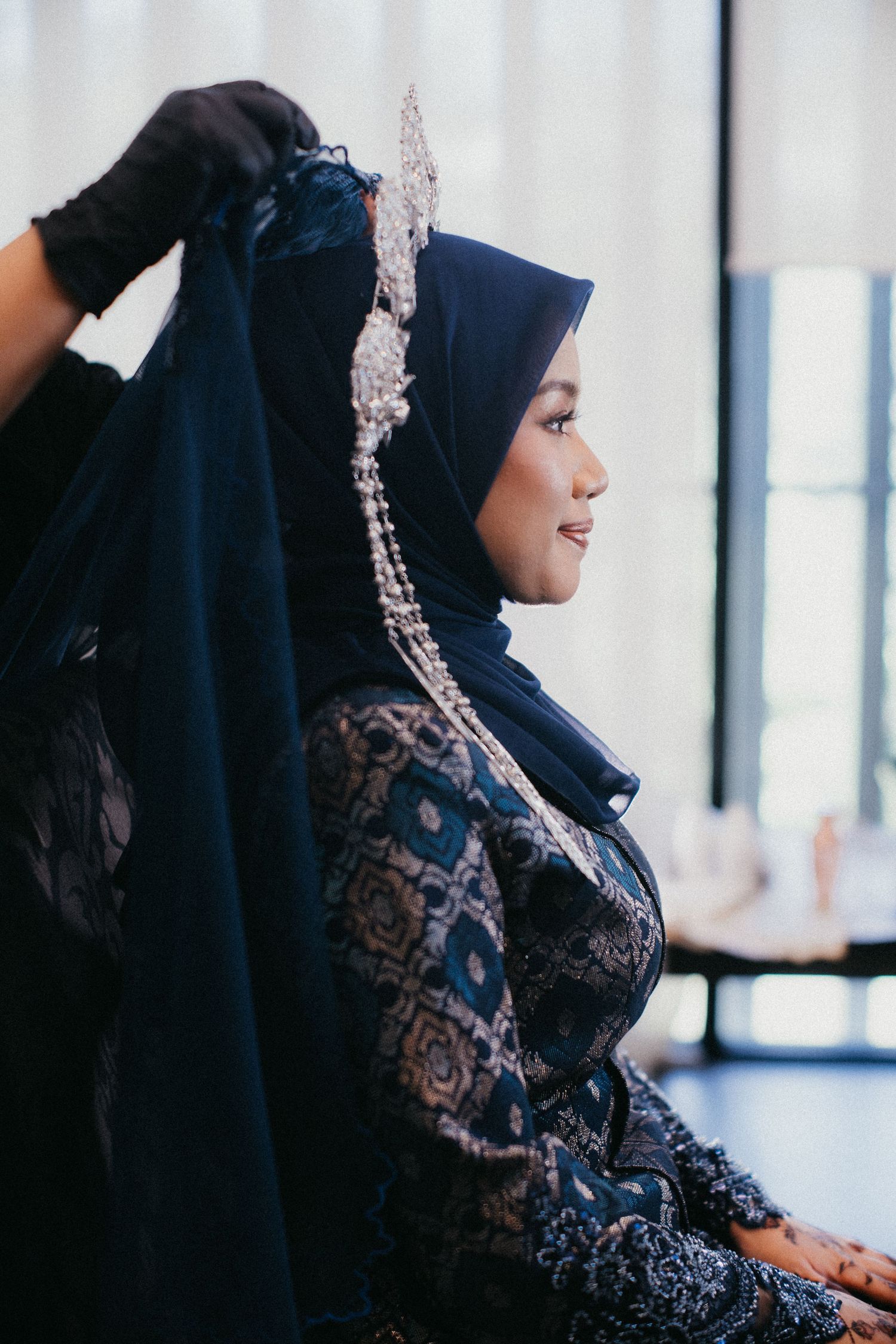 A woman wearing a dark navy hijab and lace dress sits in profile, with an elegant crystal headpiece adorning her veil.