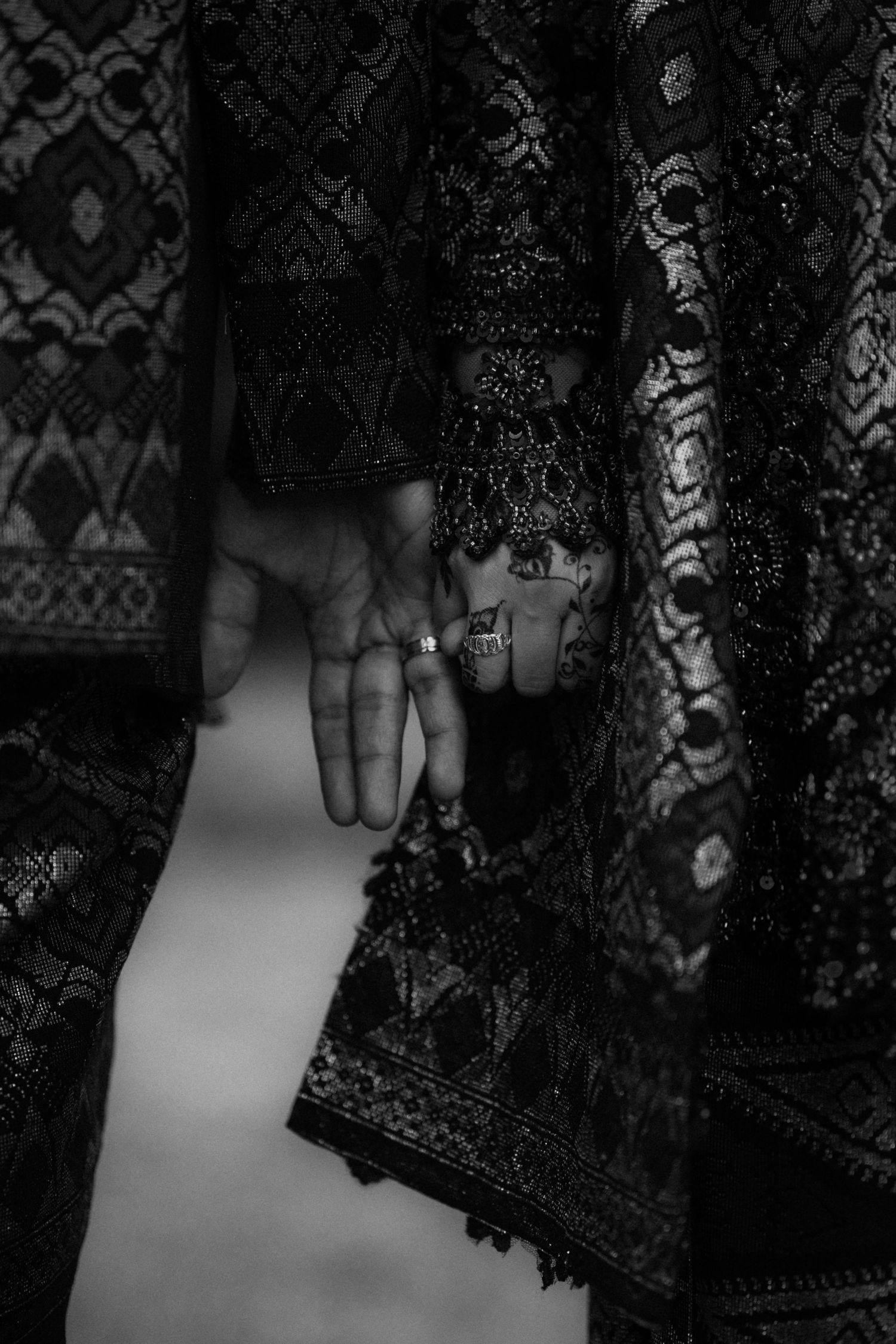 A hand adorned with rings emerges from intricate black lace fabric with ornate patterns in a dramatic monochrome shot.